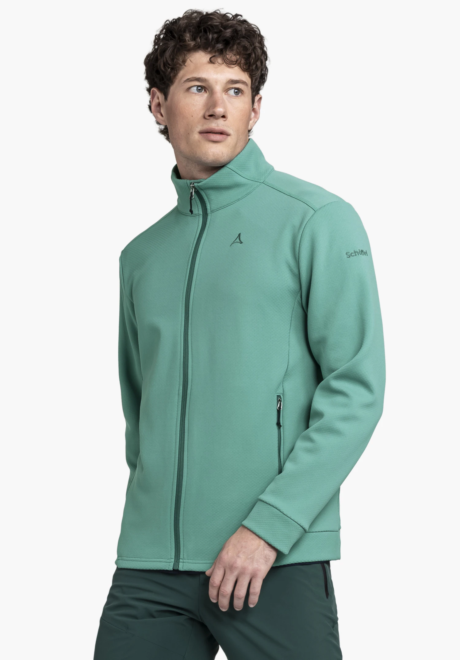Fleece Jk Style Uriezzo MNS. Ein Model trägt die Fleecejacke in Grün und steht leicht seitlich vor einem neutralen Hintergrund. Deutlich zu erkennen sind der Stehkragen, der durchgehende Reißverschluss sowie das Schöffel-Logo auf der linken Brust und am Ärmel.