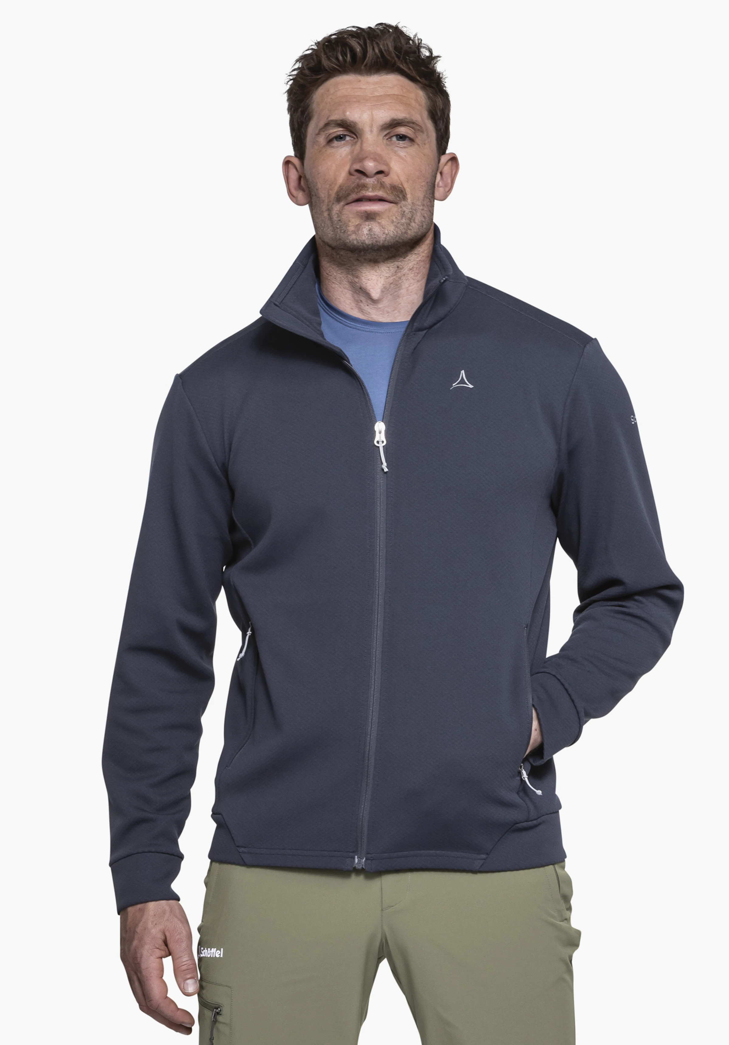 Fleece Jk Style Uriezzo MNS est porté par un mannequin se tenant de face, présentant la veste polaire bleue avec le logo Schöffel visible sur la poitrine gauche et une main dans la poche latérale, photographié devant un fond neutre.