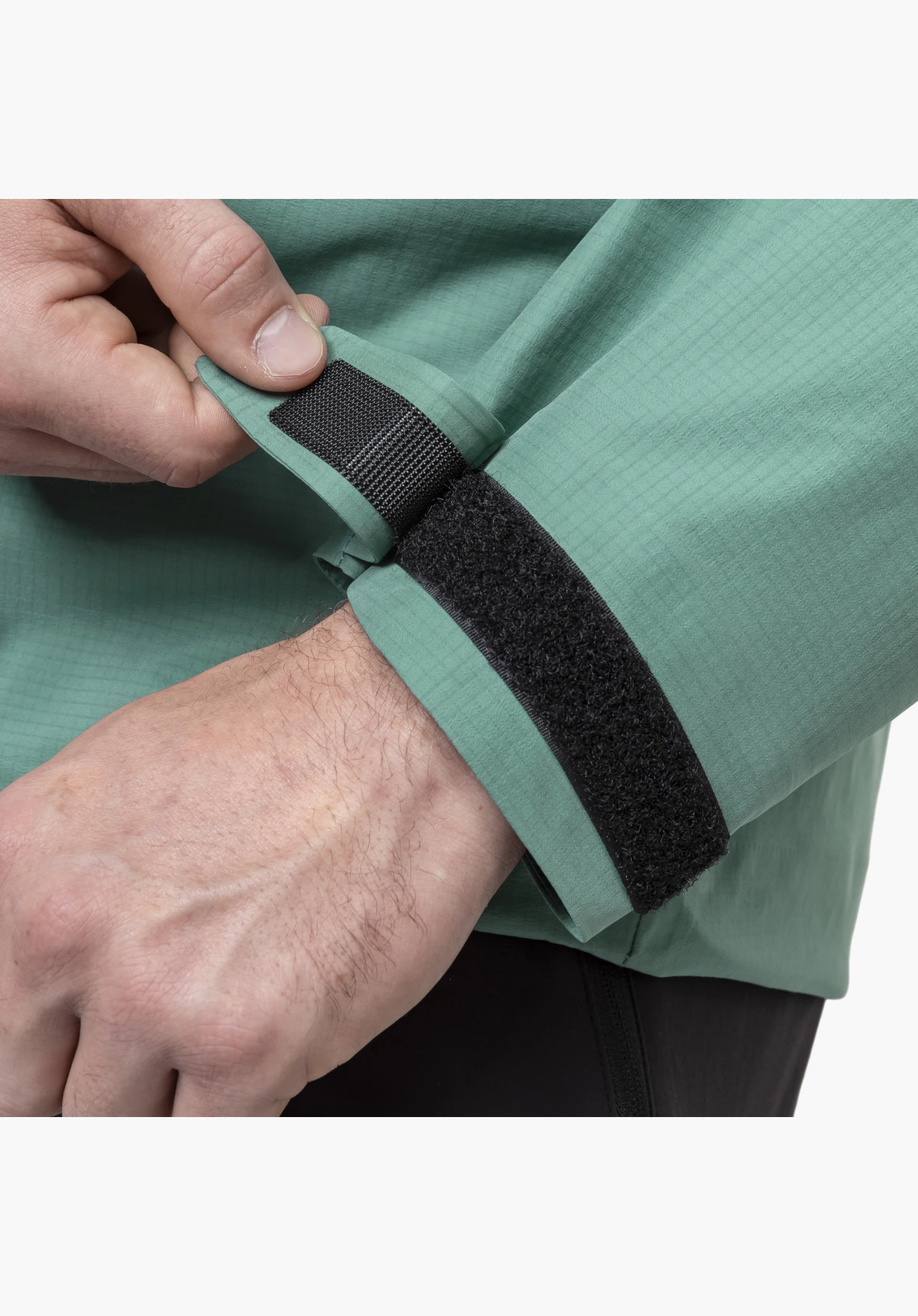 Jacket Style Blaueis MNS montre le poignet de la manche verte avec une fine texture de tissu, où une main ajuste la fermeture autoagrippante noire, gros plan sur un fond blanc/neutre.