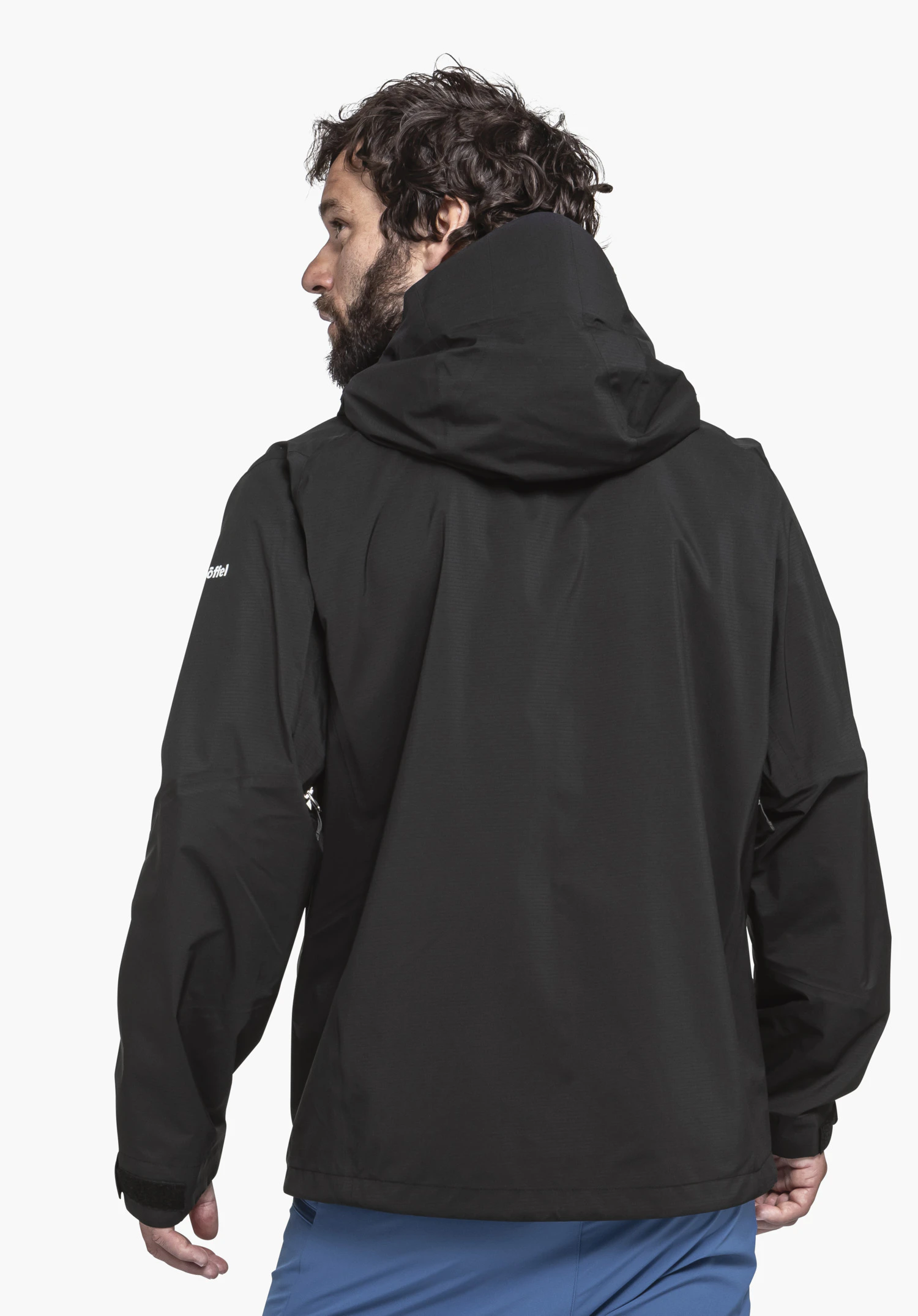Jacket Style Blaueis MNS schwarze Regenjacke, getragen von Person, dreht den Kopf nach links; Kapuze hoch, weißer Schriftzug "Schöffel" am Ärmel; steht vor hellgrauem Hintergrund, trägt hellblaue Hose.