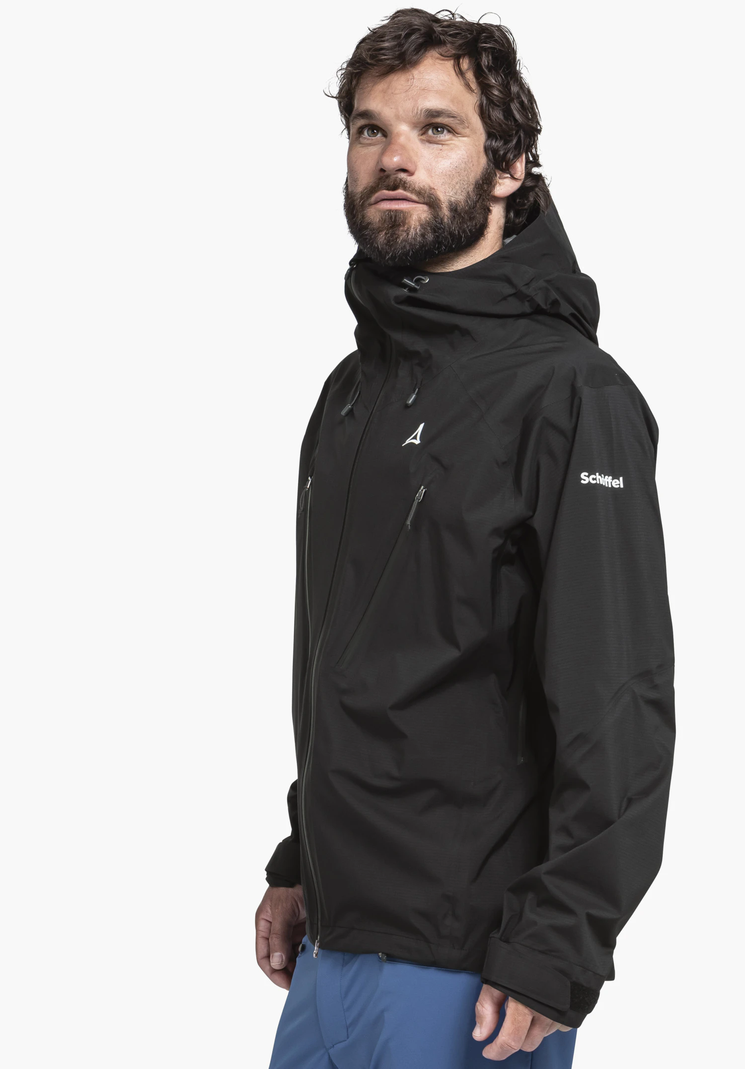 Jacket Style Blaueis MNS Schwarze Kapuzenjacke (Schöffel-Logo) getragen von einem Mann, der nach oben blickt; blaue Hose; vor hellweißem Hintergrund. Text auf Ärmel: "Schöffel".