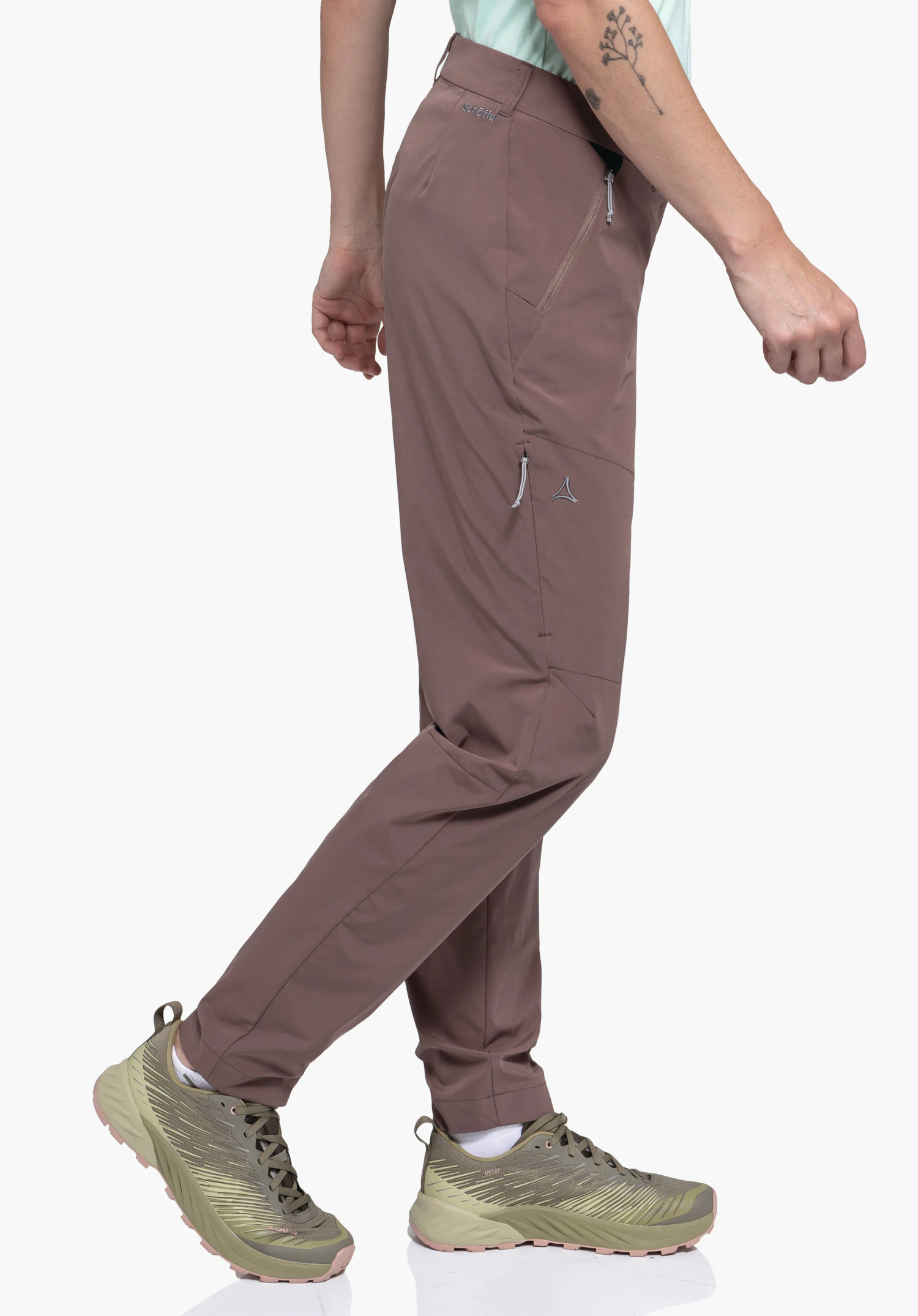 CIRC Pants Style Smue WMS portée par un mannequin se tenant de profil dans une posture de marche. Le pantalon marron présente des poches zippées pratiques ainsi que le logo Schöffel sur la cuisse et la taille, photographié sur un fond neutre.