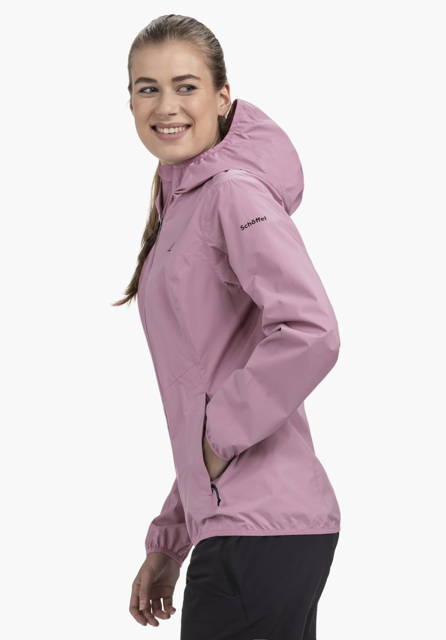 Jacket Style Migandi WMS sur un mannequin qui se tient de profil et sourit à la caméra. Elle porte la veste rose avec capuche et a une main posée dans la poche latérale, avec le Schöffel-Logo bien visible sur le haut du bras gauche, présenté sur un fond neutre.