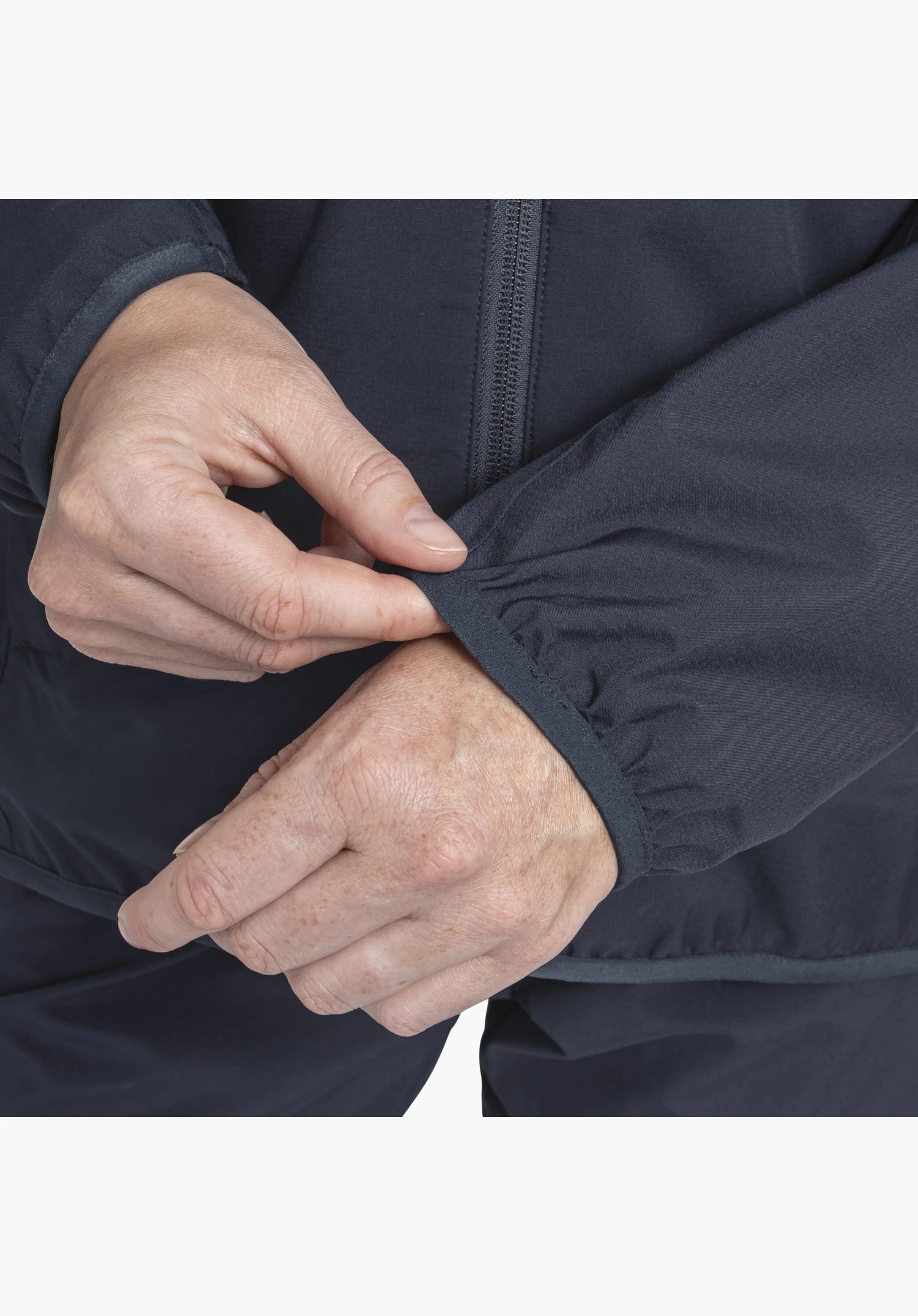 Jacket Style Migandi WMS montre le poignet élastique de la veste bleue ajusté par deux mains, ainsi qu'une fermeture zippée avant partiellement visible en gros plan sur fond blanc/neutre.