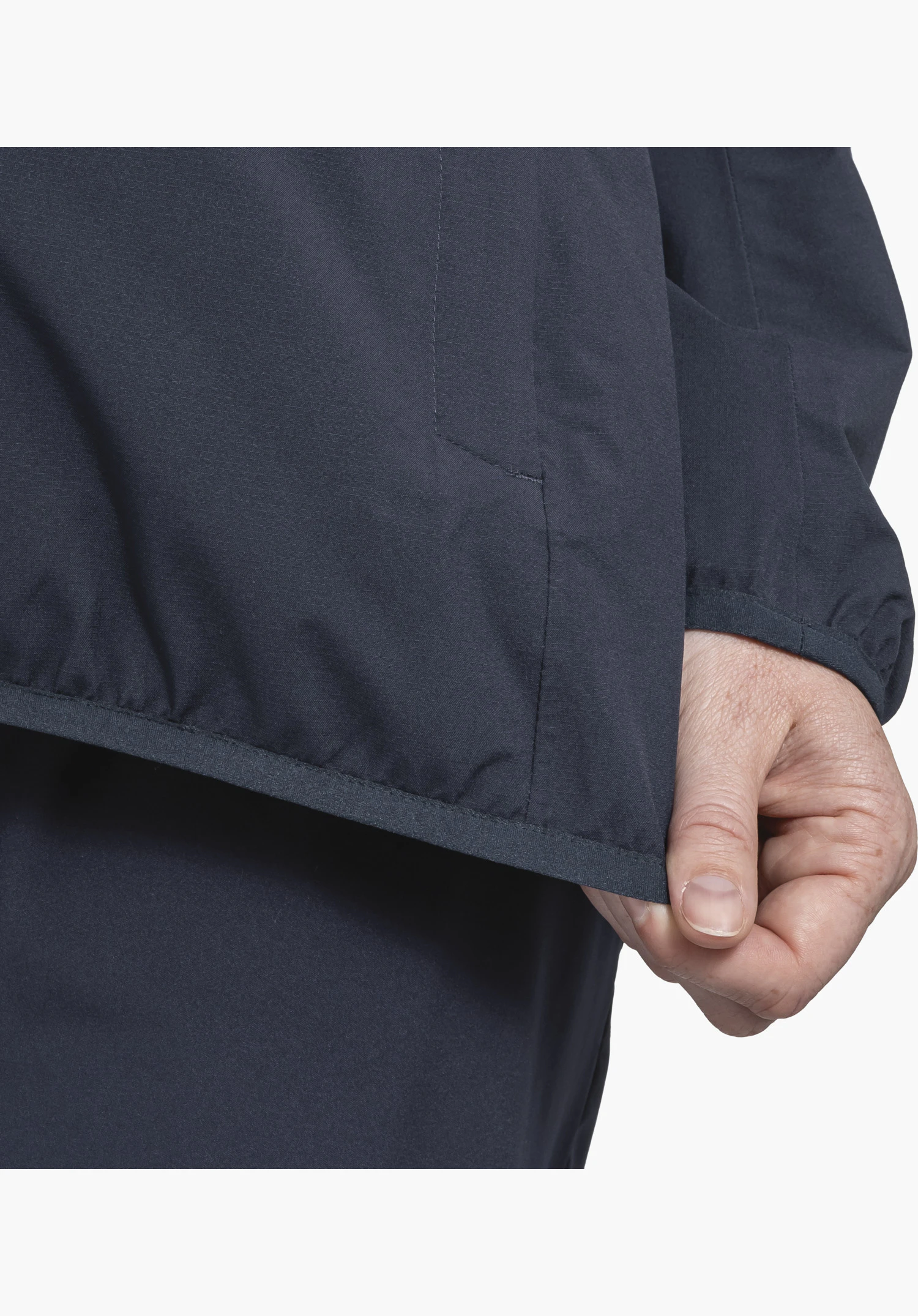 Jacket Style Migandi WMS montre le tissu bleu avec une bordure élastique sur l'ourlet inférieur et le poignet, tenu par une main, en gros plan sur fond blanc/neutre.