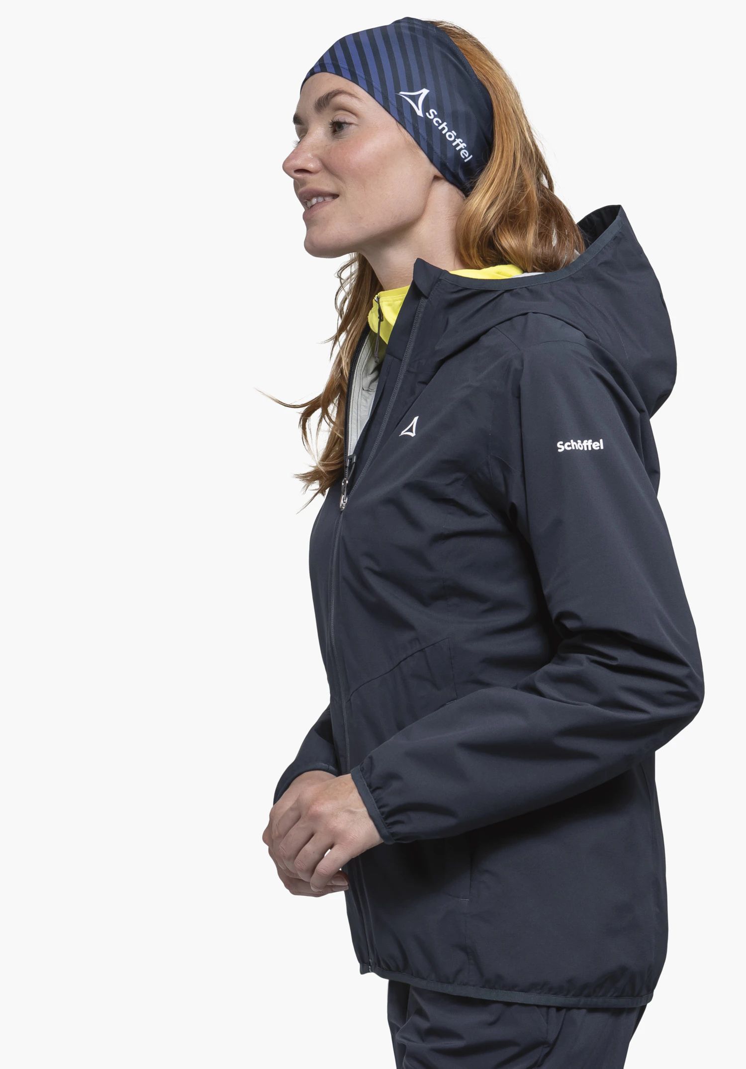 Jacket Style Migandi WMS sur un mannequin femme qui se tient de profil et porte la veste bleue. Le logo Schöffel blanc est bien visible sur la poitrine, la manche ainsi que sur le bandeau. La photo présente la vue portée devant un fond neutre.