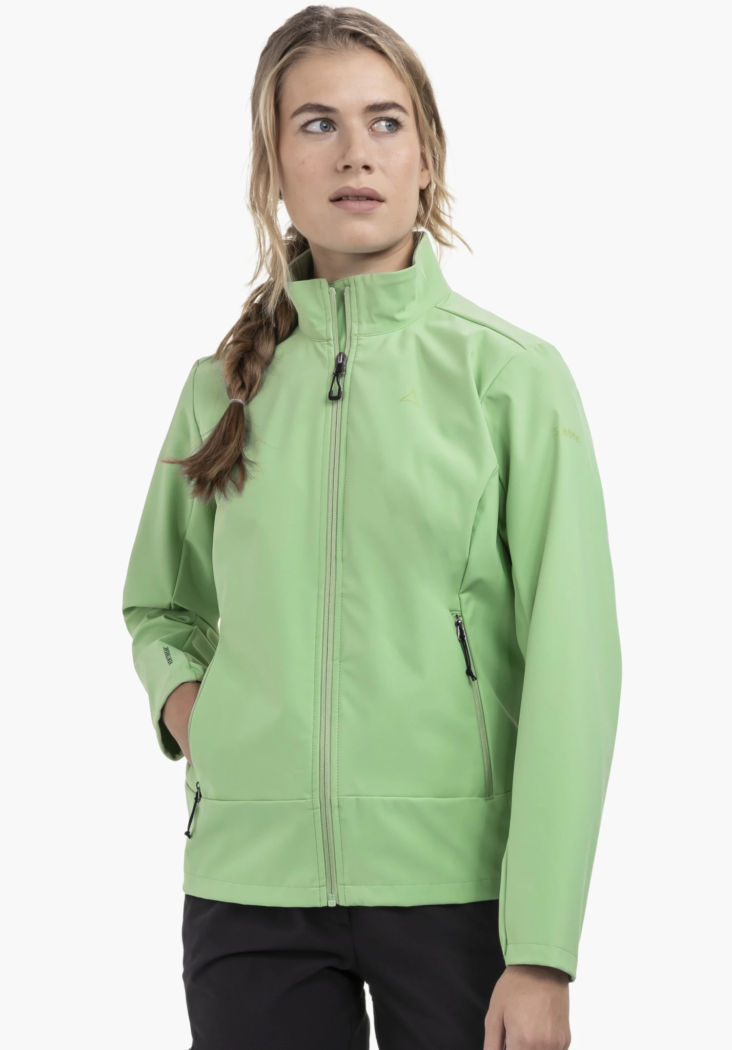 Softshell Jk Style Mirusha WMS in der Farbe Grün. Das Model steht leicht schräg mit einer Hand in der Seitentasche und trägt die hochgeschlossene Jacke, auf deren linker Brust das Schöffel-Logo dezent zu sehen ist. Präsentiert wird das Outfit vor einem neutralen Hintergrund.