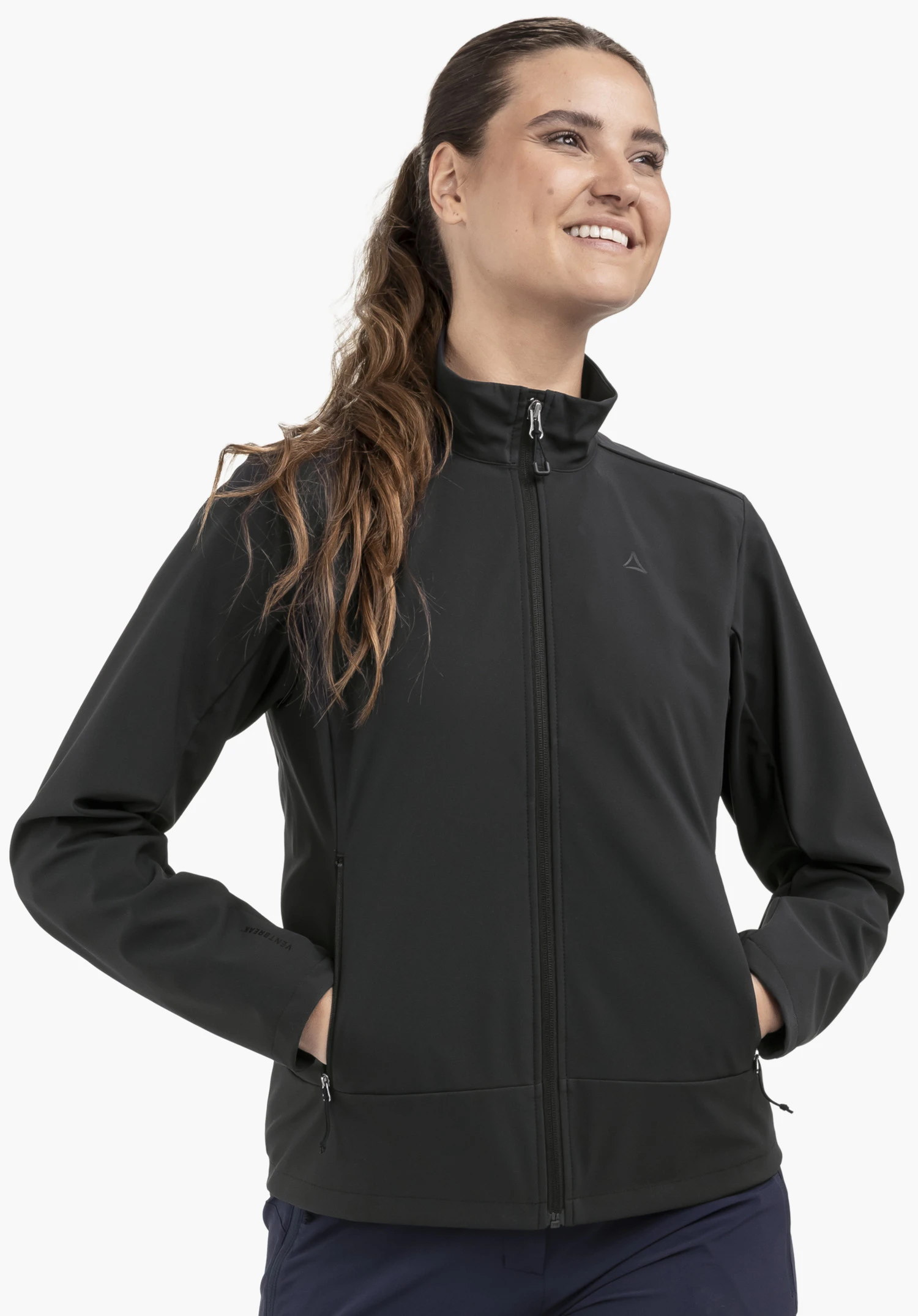 Softshell Jk Style Mirusha WMS sur un mannequin qui se tient de face et regarde en souriant sur le côté. Les mains reposent dans les poches de la veste noire, qui affiche le logo Schöffel visible sur la poitrine gauche, photographiée devant un fond neutre.
