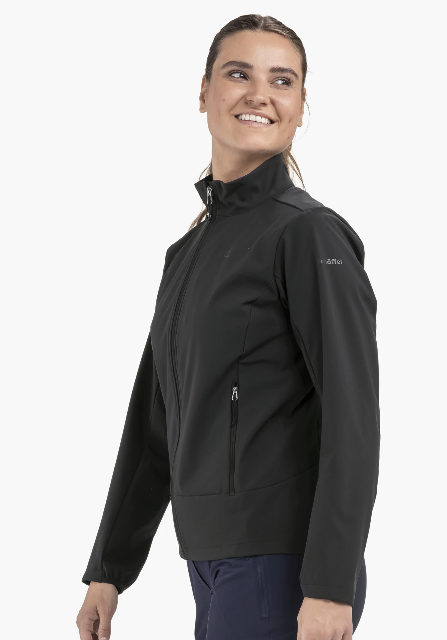 Softshell Jk Style Mirusha WMS in zwart voor een neutrale achtergrond. Het model staat zijwaarts en kijkt met een vriendelijke glimlach over haar schouder. Het subtiele Schöffel-logo op de bovenarm is duidelijk zichtbaar.