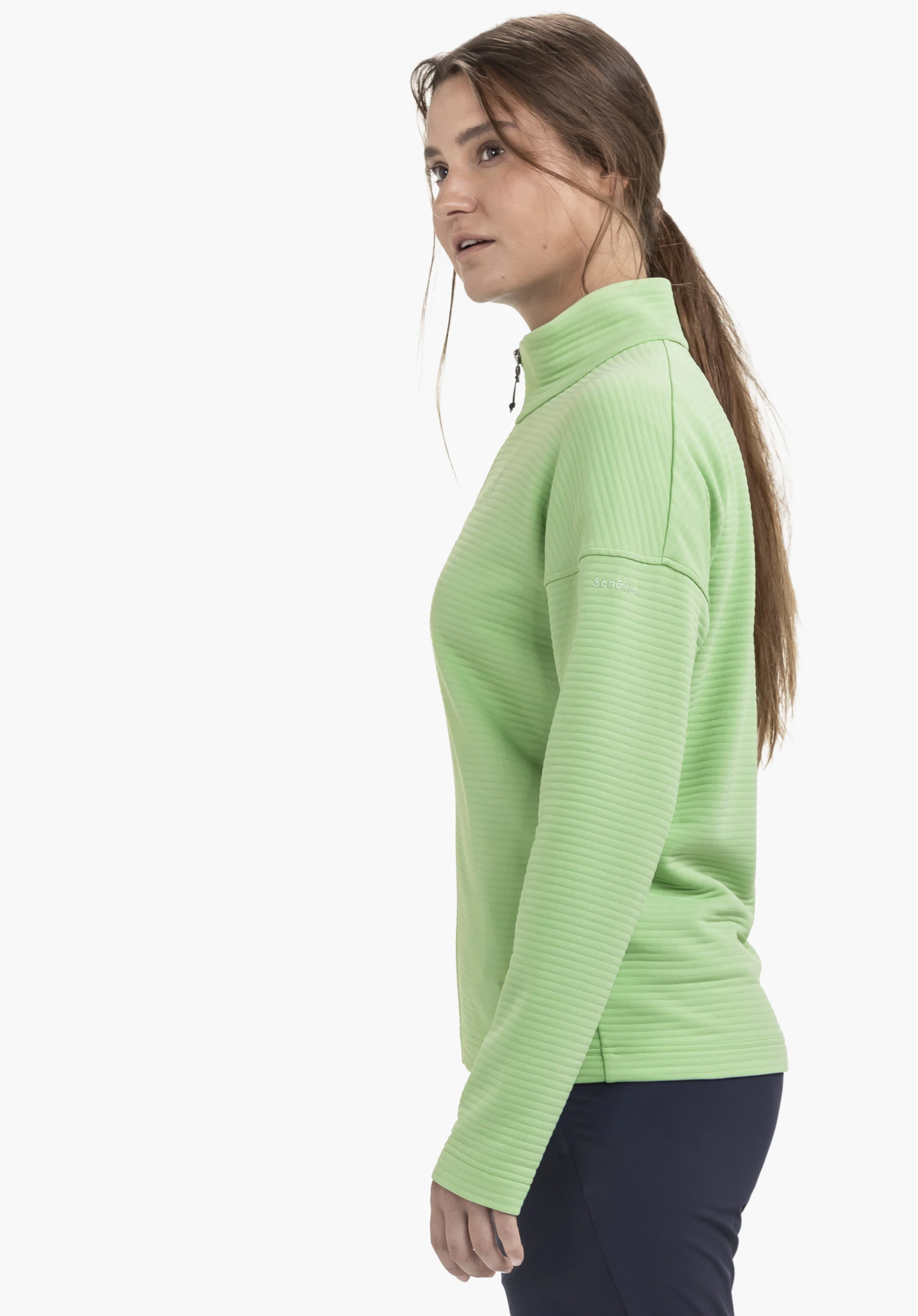 CIRC Fleece Jk Style Smue WMS: Eine Frau steht seitlich zum Betrachter vor einem neutralen Hintergrund und präsentiert die Fleecejacke in Grün. Das dezent platzierte Schöffel-Logo am Ärmel unterstreicht das minimalistische Design des Modells.