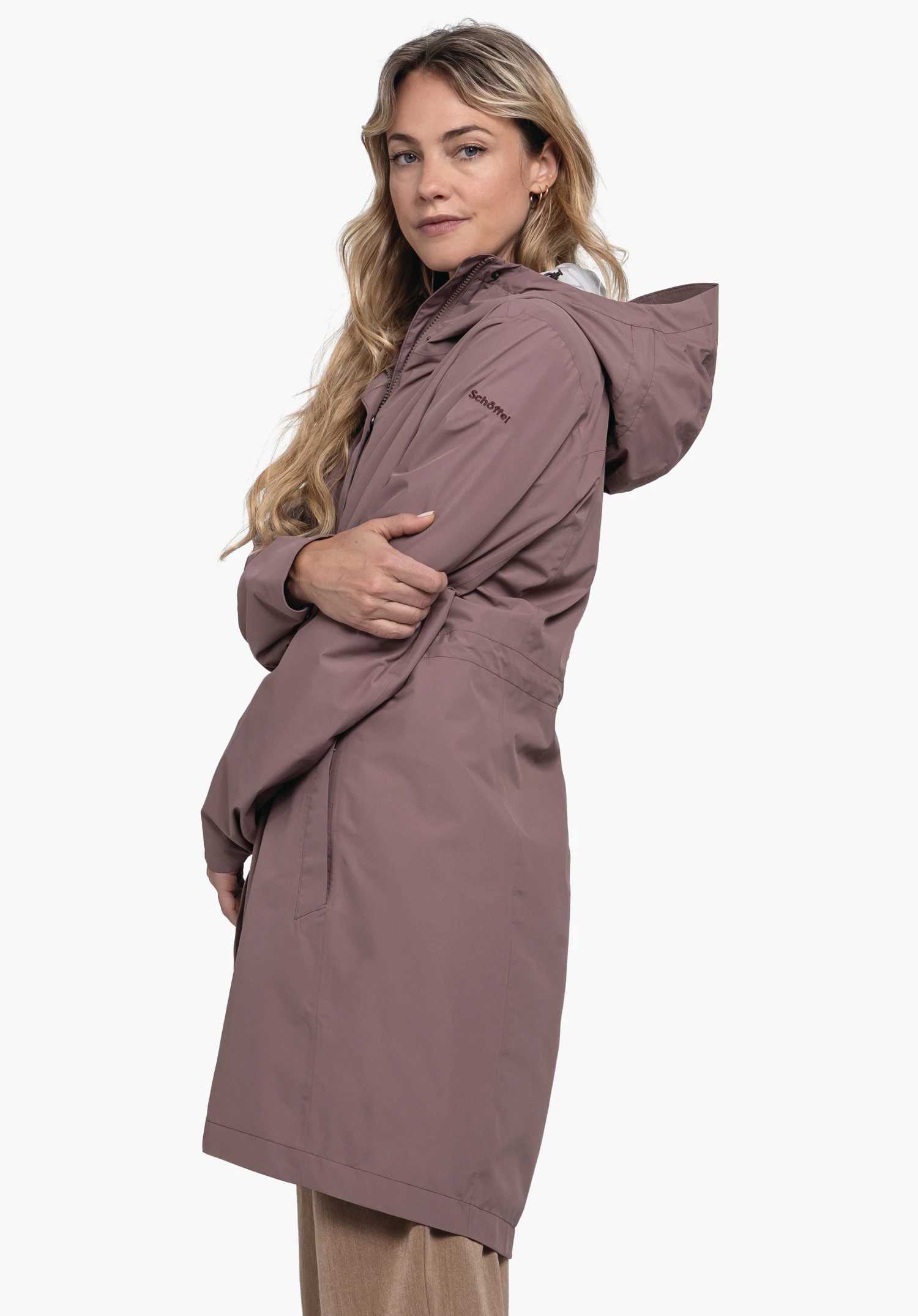 Coat Style Bohorok WMS. The model is standing sideways in front of a neutral background, showcasing the coat in a stylish brown. She looks directly into the camera, while the Schöffel logo is subtly visible on the sleeve.