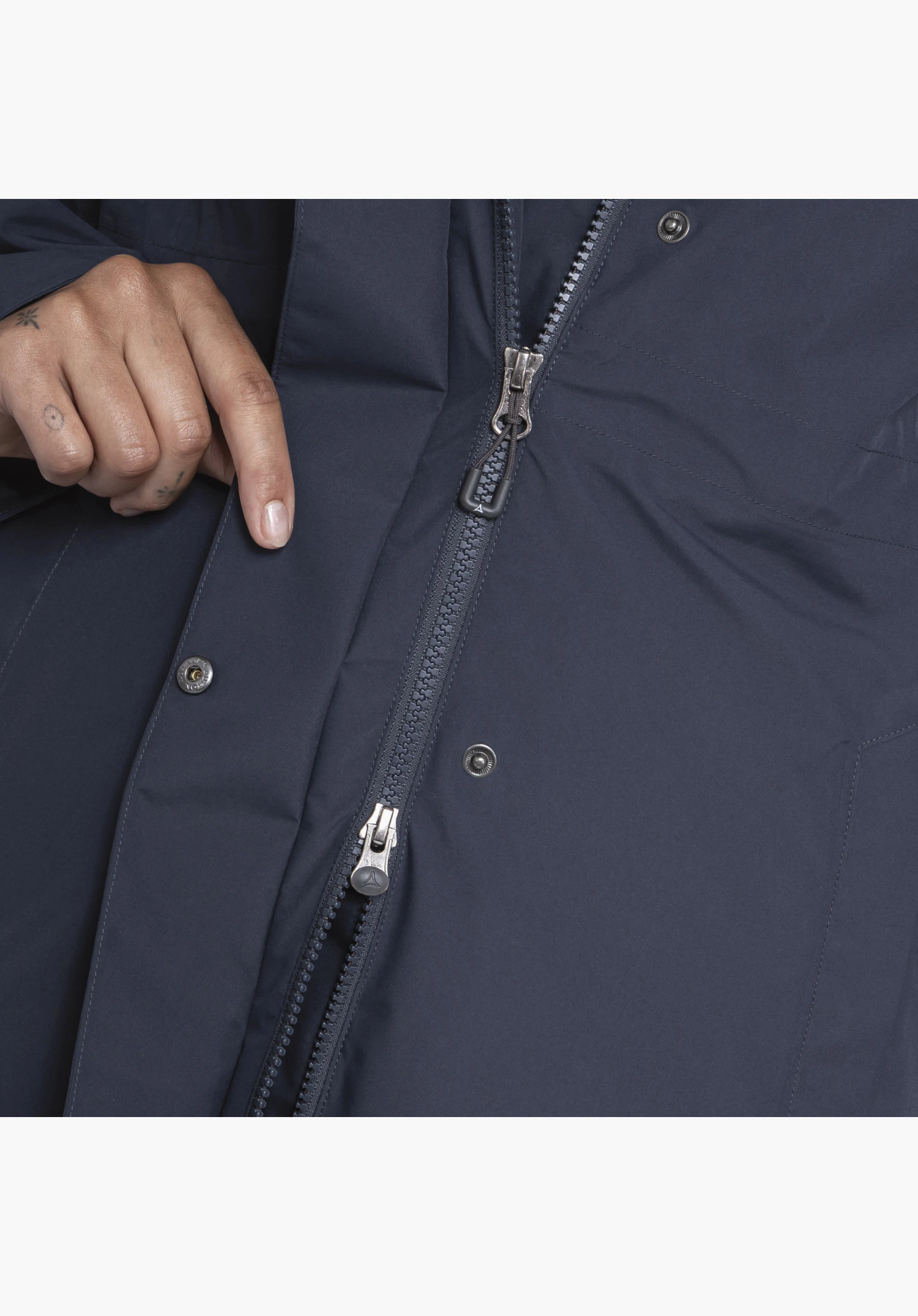 Coat Style Bohorok WMS shows in detail the two-way zip on the blue fabric. The lower puller clearly features the Schöffel logo, whilst a hand slightly lifts the placket with its metal snap fasteners. Close-up against a white/neutral background.