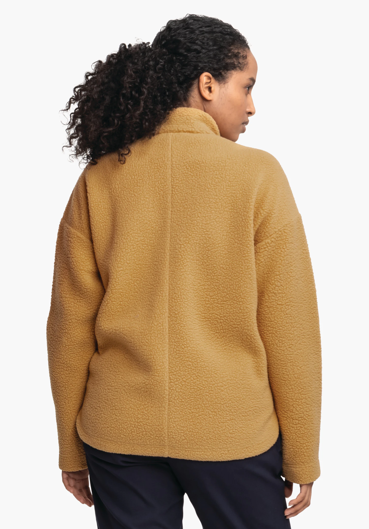 CIRC Fleece Jk Style Purga WMS Fleecejacke: Frau steht mit dem Rücken zur Kamera, trägt eine dunkel gelbe Teddy-Fleecejacke, schwarzes lockiges Haar zu Pferdeschwanz, dunkel blaue Hose, weißer Hintergrund.