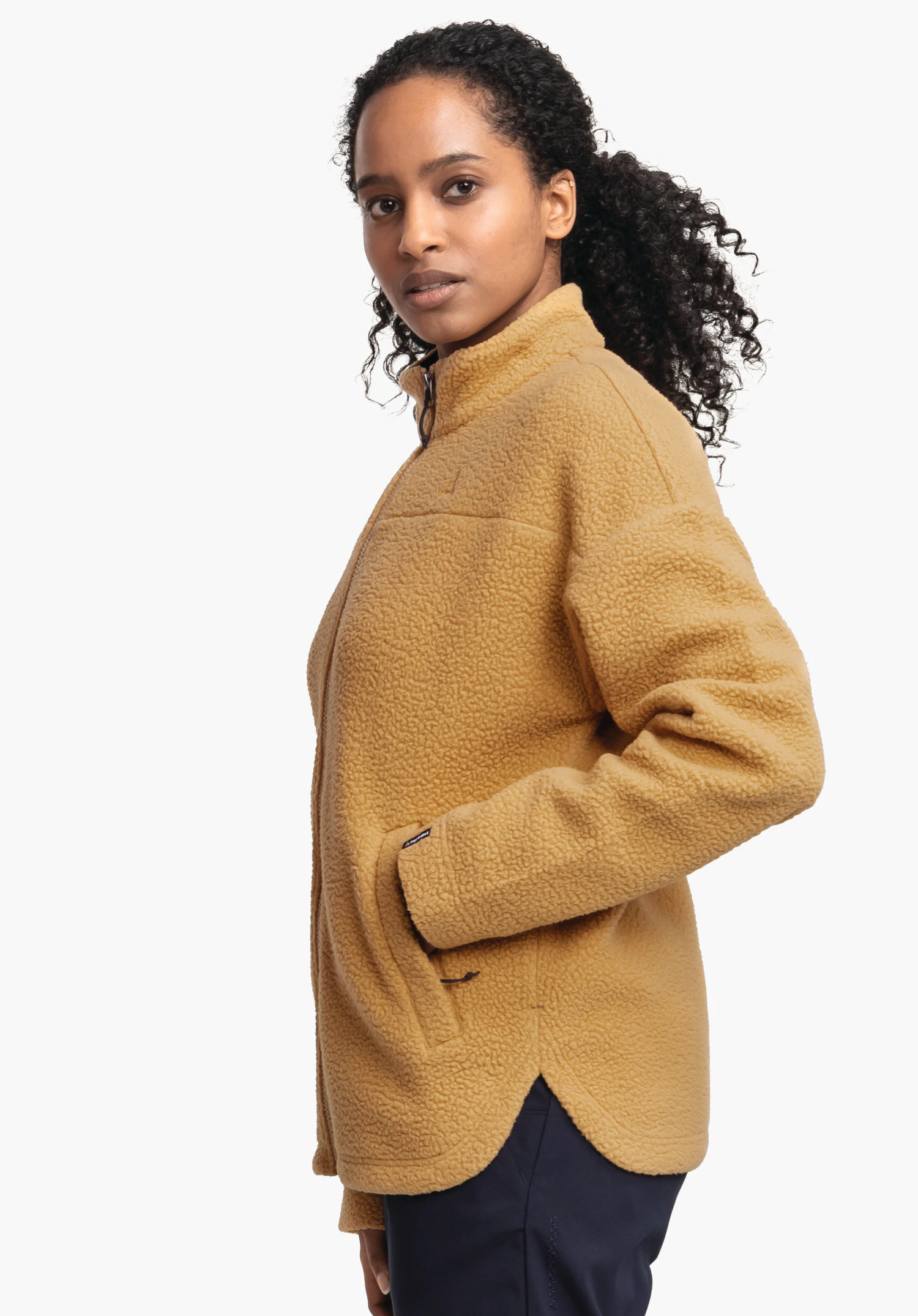 CIRC Fleece Jk Style Purga WMS dunkelgelbe Fleecejacke, getragen von Person mit lockigen Haaren; Jacke geschlossen, rechte Hand in Seitentasche, Person steht seitlich vor weißem Hintergrund, dunkelblaue Hose sichtbar.