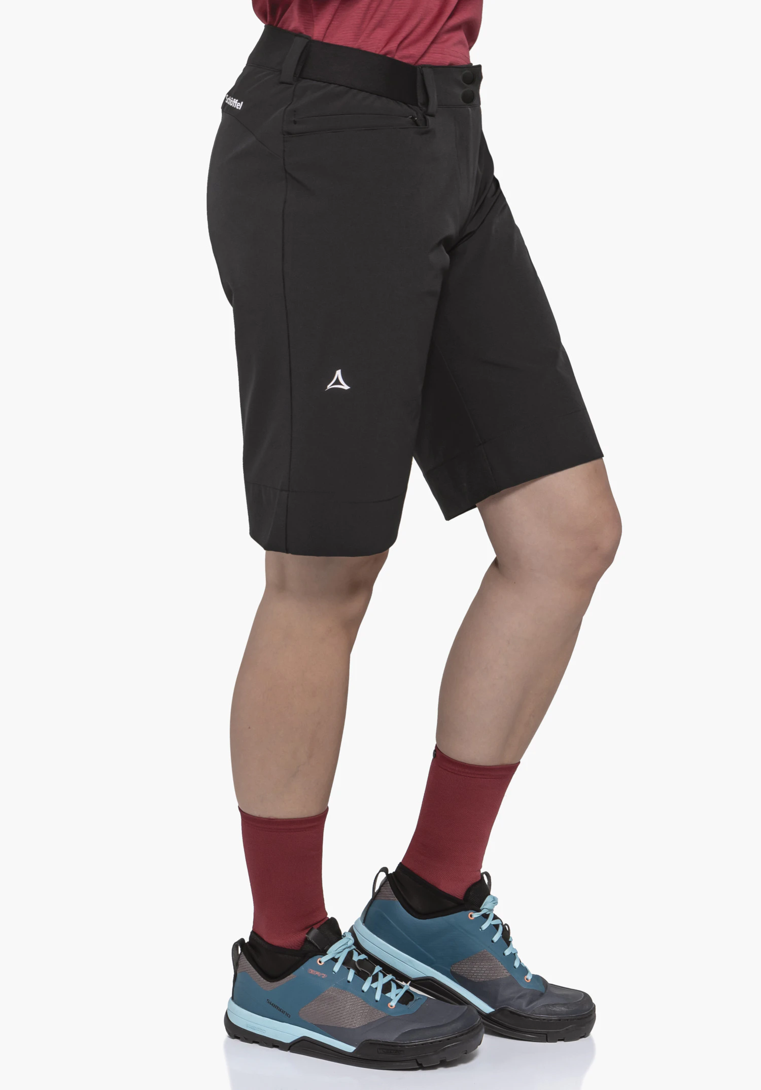 Shorts Style Keitele WMS zwarte shorts, zijwaarts gedragen door een model, net boven de knie; Schöffel-logo op het bovenbeen; gecombineerd met rode sokken en blauwe schoenen met lichtblauwe accenten. Tekst: "Schöffel".