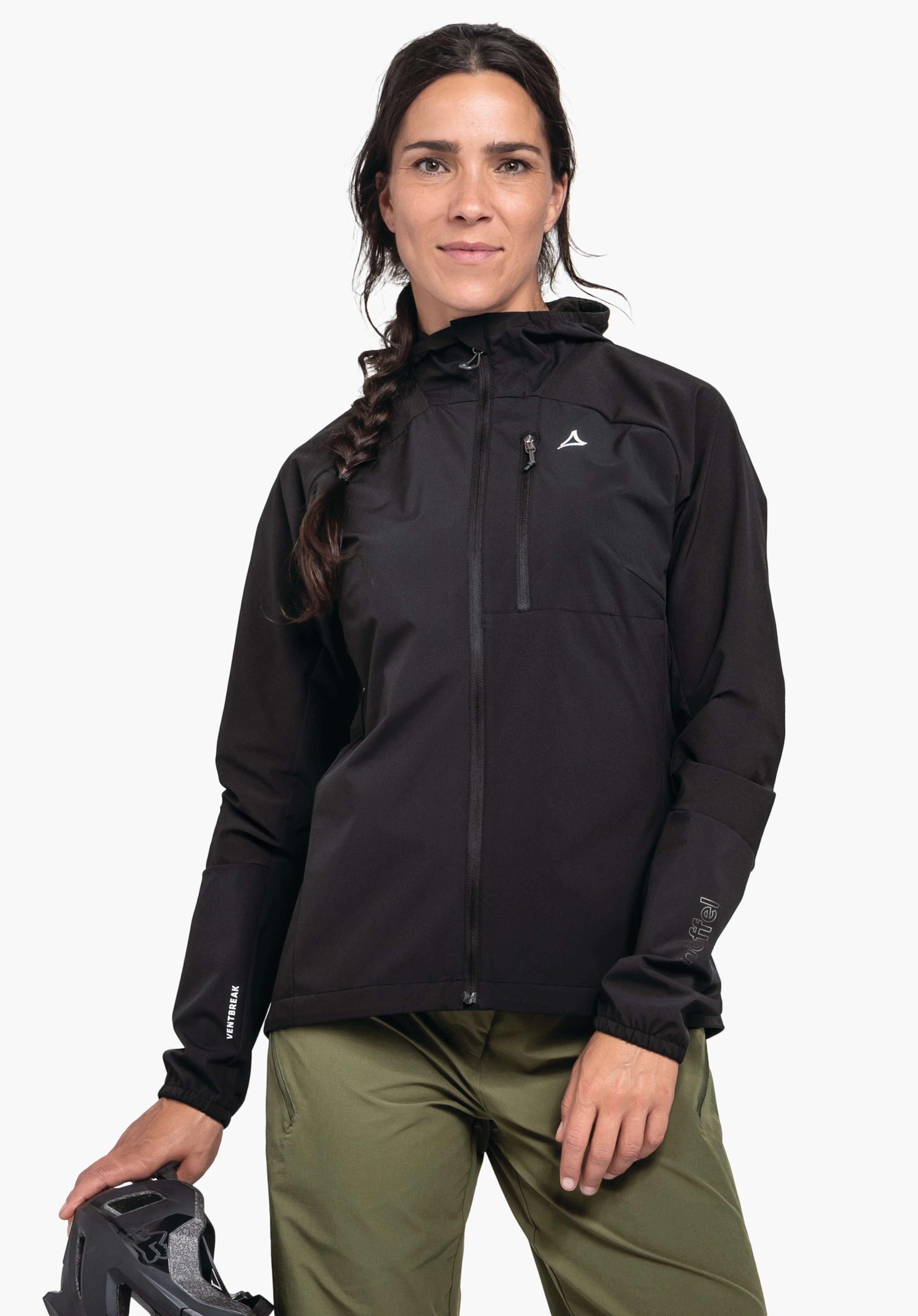 Hybrid Jk Style Mamry WMS in der Farbe Schwarz. Das Model steht frontal, blickt in die Kamera und präsentiert die geschlossene Jacke mit sichtbarem Schöffel-Logo auf der linken Brust. In der rechten Hand hält sie einen Fahrradhelm, aufgenommen vor einem neutralen Hintergrund.