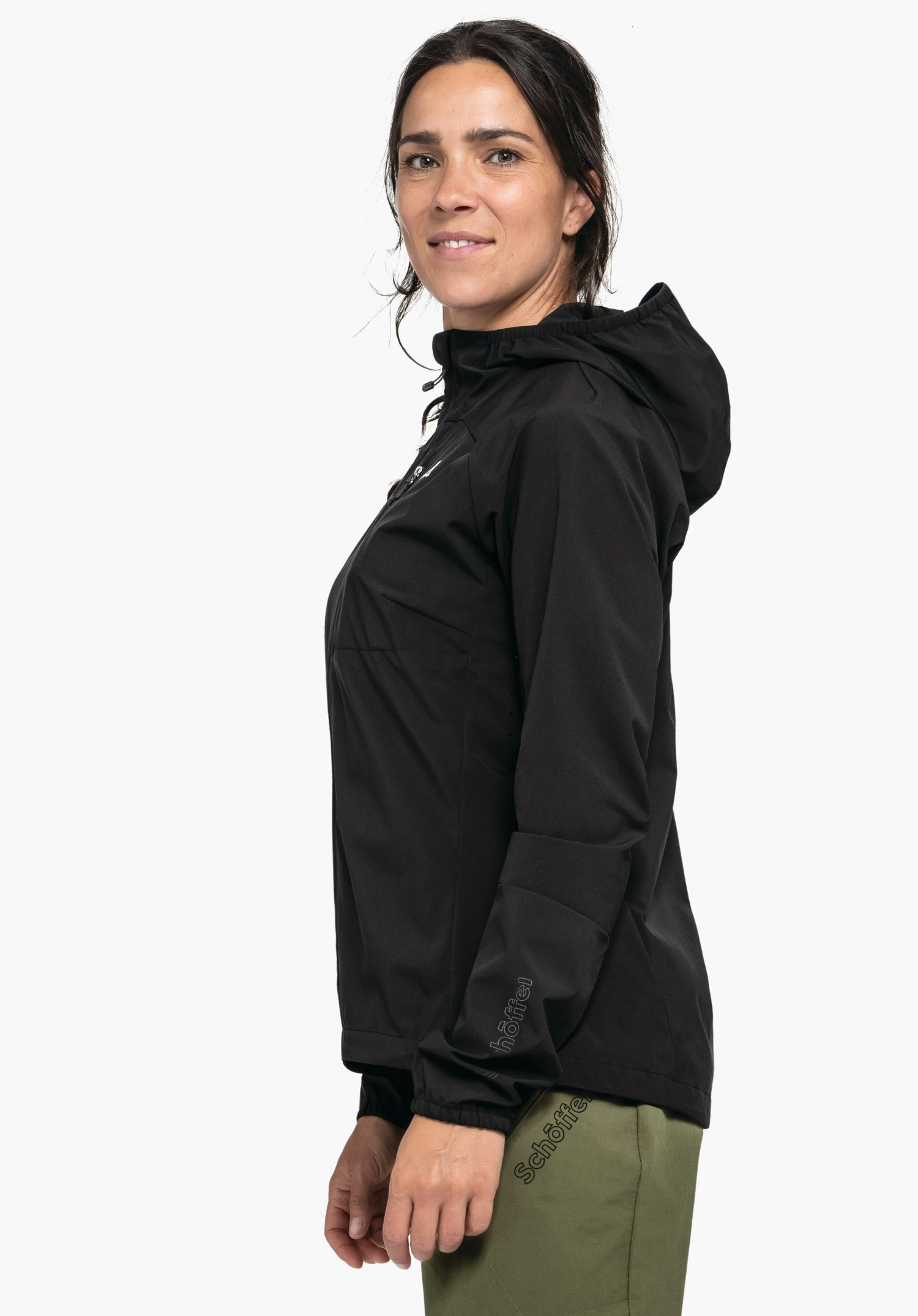 Hybrid Jk Style Mamry WMS en noir. Une femme sportive se tient debout de profil devant un fond neutre et porte la veste. Le logo Schöffel discret est visible sur le bas de la manche et au niveau de la poitrine.