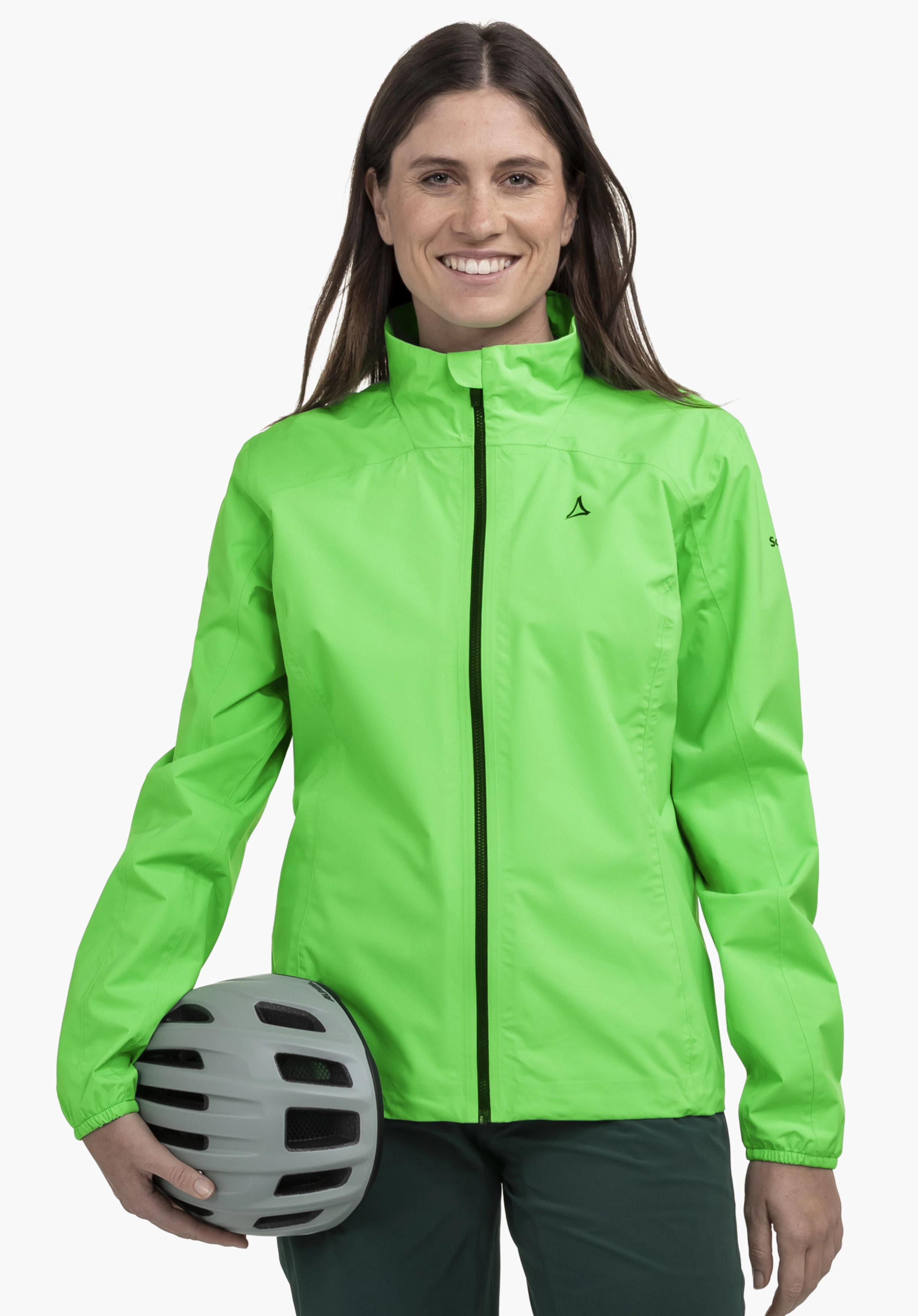 Jacket Style Lurbek WMS an einem Model, das frontal steht und in die Kamera lächelt. Sie trägt die grüne Jacke mit dem sichtbaren Schöffel-Logo auf der linken Brust und hält einen Fahrradhelm im rechten Arm vor einem neutralen Hintergrund.