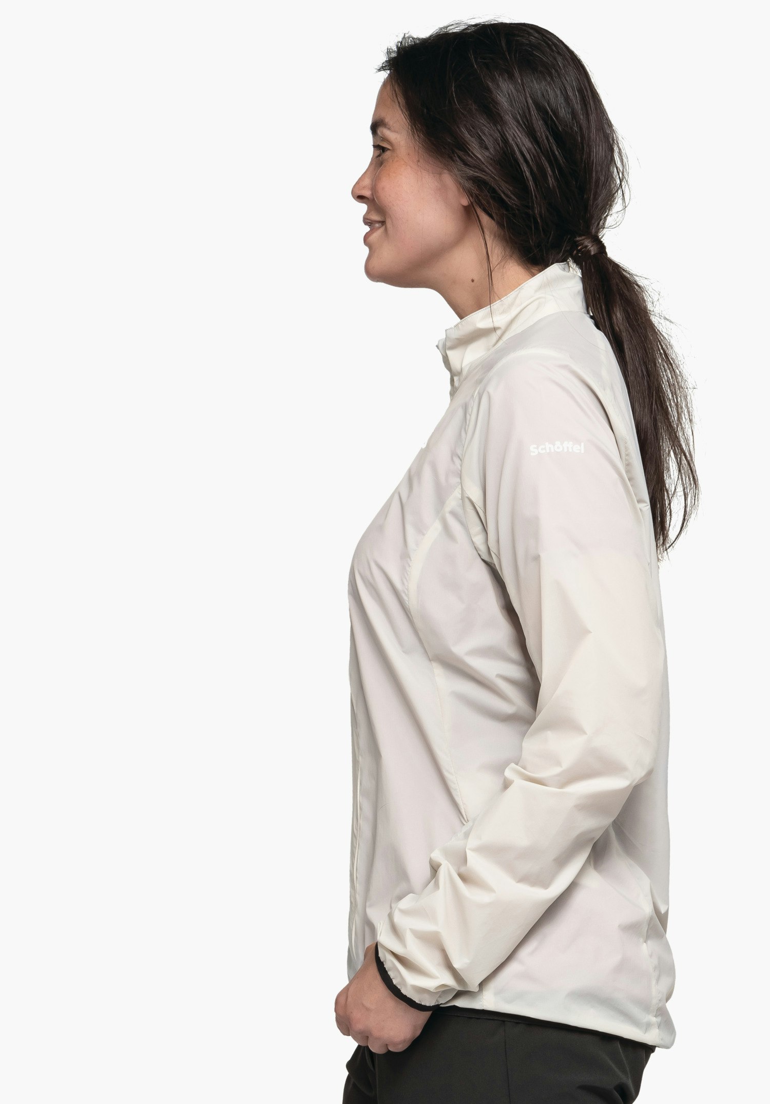Jacket Style Cannobio WMS woman stands side-on in profile wearing a light white jacket with the lettering "Schöffel", smiles slightly and has her hands in the pockets in front of a plain white background.