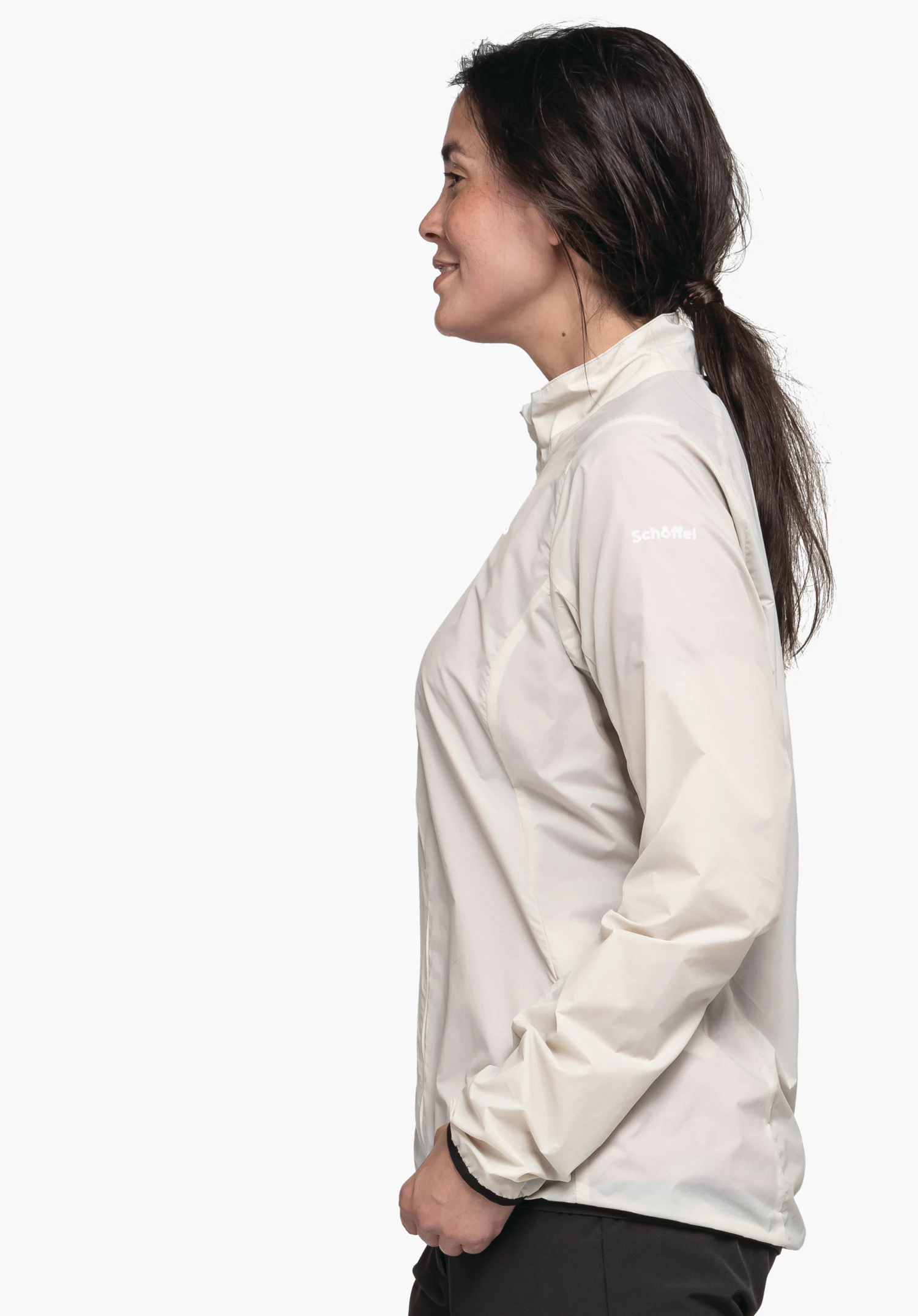Jacket Style Cannobio WMS femme debout de profil, porte une veste blanc clair avec l’inscription "Schöffel", sourit légèrement et a les mains dans les poches devant fond blanc sobre.