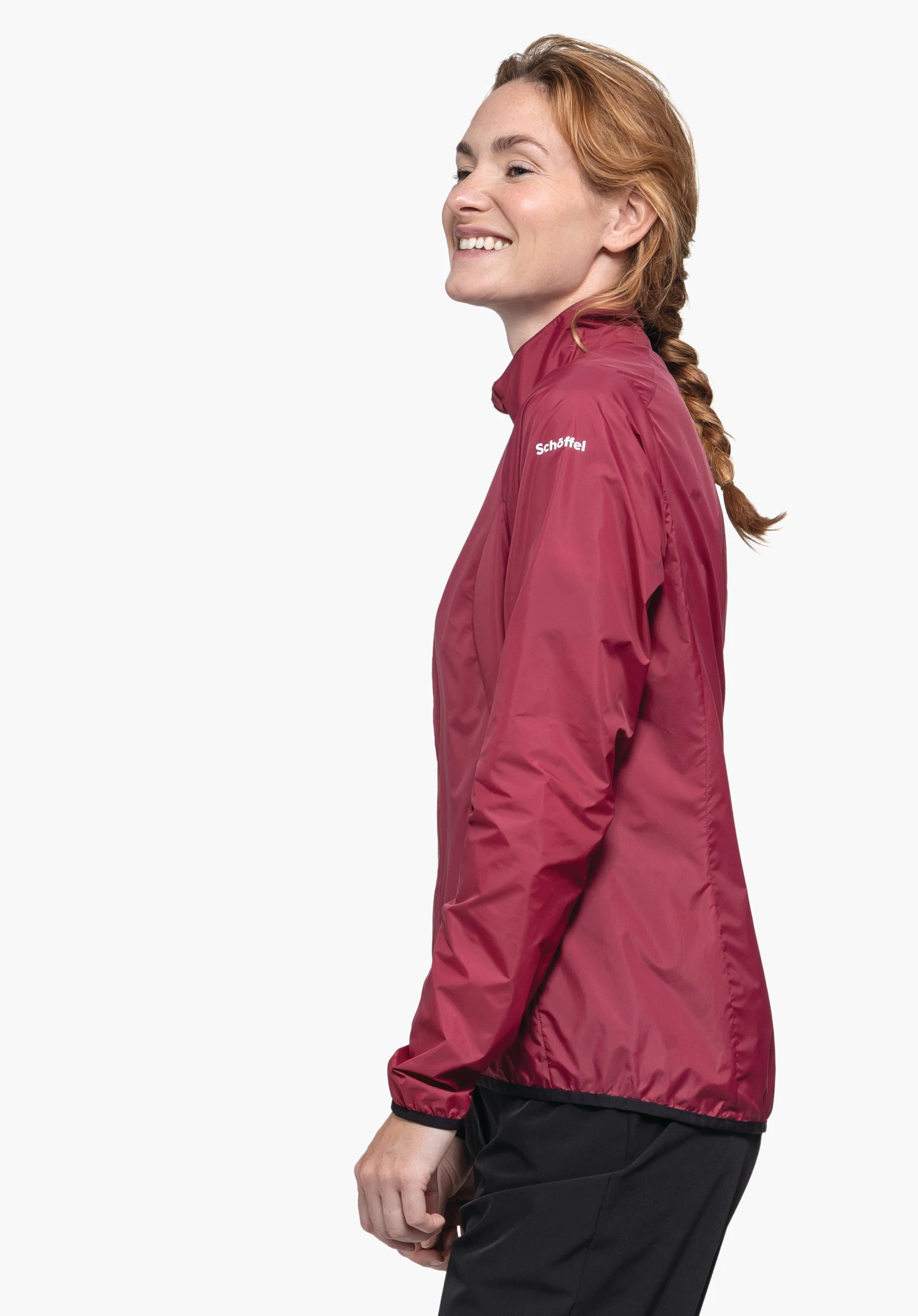 Jacket Style Cannobio WMS Frau trägt dunkelrote Jacke, lächelt und schaut seitlich; braune geflochtene Haare, schwarze Hose vor weißem Hintergrund. Text: "Schöffel".