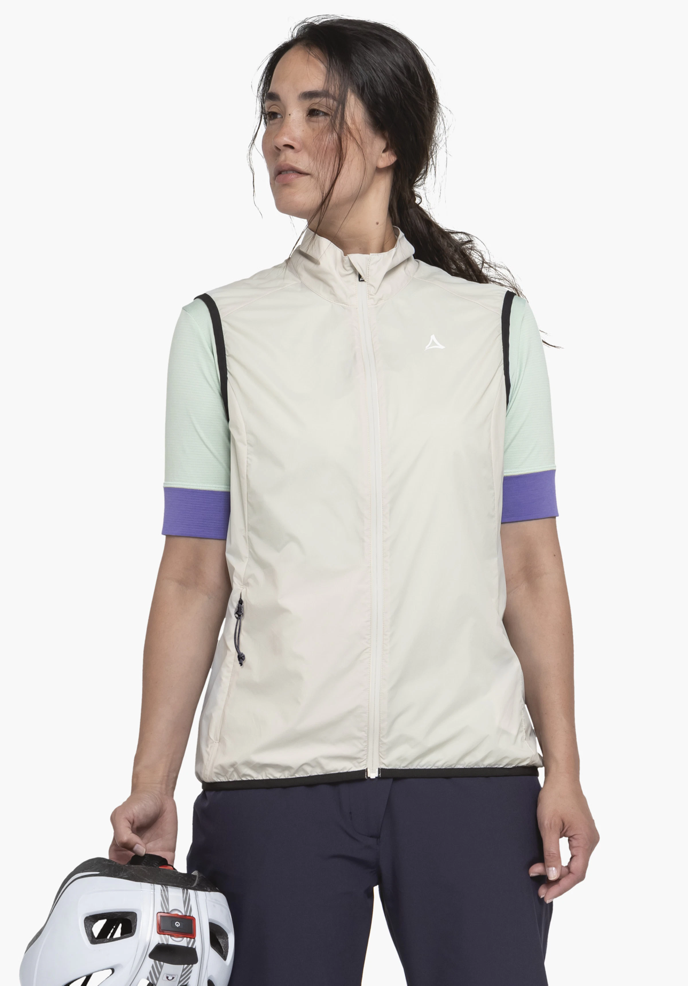 Vest Style Cannobio WMS in der Farbe Natur. Das Model steht frontal, richtet den Blick zur Seite und hält einen Fahrradhelm in der Hand. Zu sehen ist die geschlossene Weste mit einem weißen Schöffel-Logo auf der Brust. Präsentiert vor einem neutralen Hintergrund.