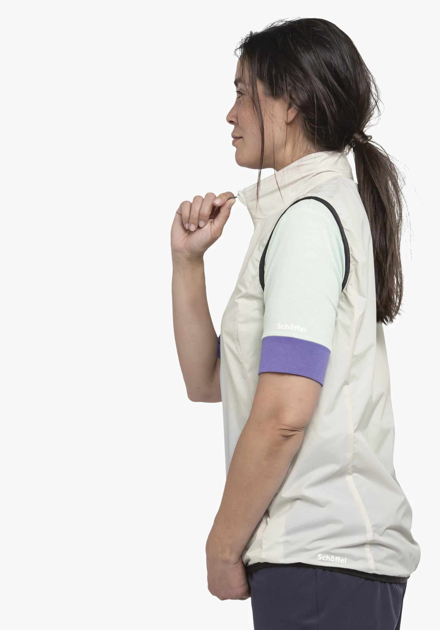 Vest Style Cannobio WMS in der Farbe Natur, präsentiert in einer seitlichen Ansicht vor einem neutralen Hintergrund. Die Sportlerin hält den Kragen der Weste, wobei das dezente Schöffel-Logo am unteren Saum sowie ein weiteres Logo am Shirt-Ärmel gut sichtbar sind.