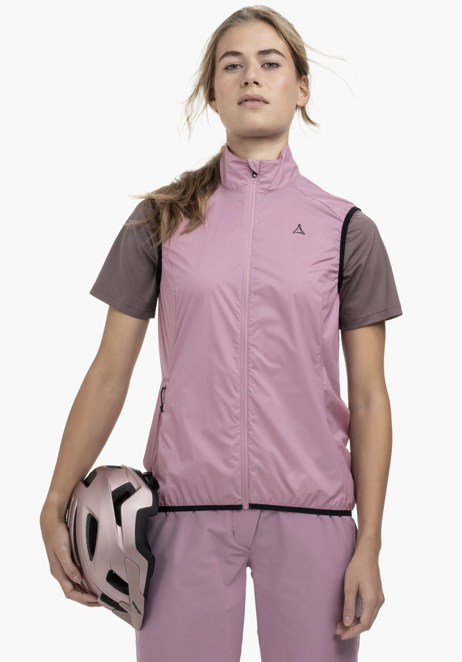 Vest Style Cannobio WMS portée par un mannequin debout de face, tenant un casque de vélo sous le bras droit. Le gilet rose présente une fermeture éclair intégrale et le logo Schöffel sur la poitrine gauche, photographié sur un fond neutre.