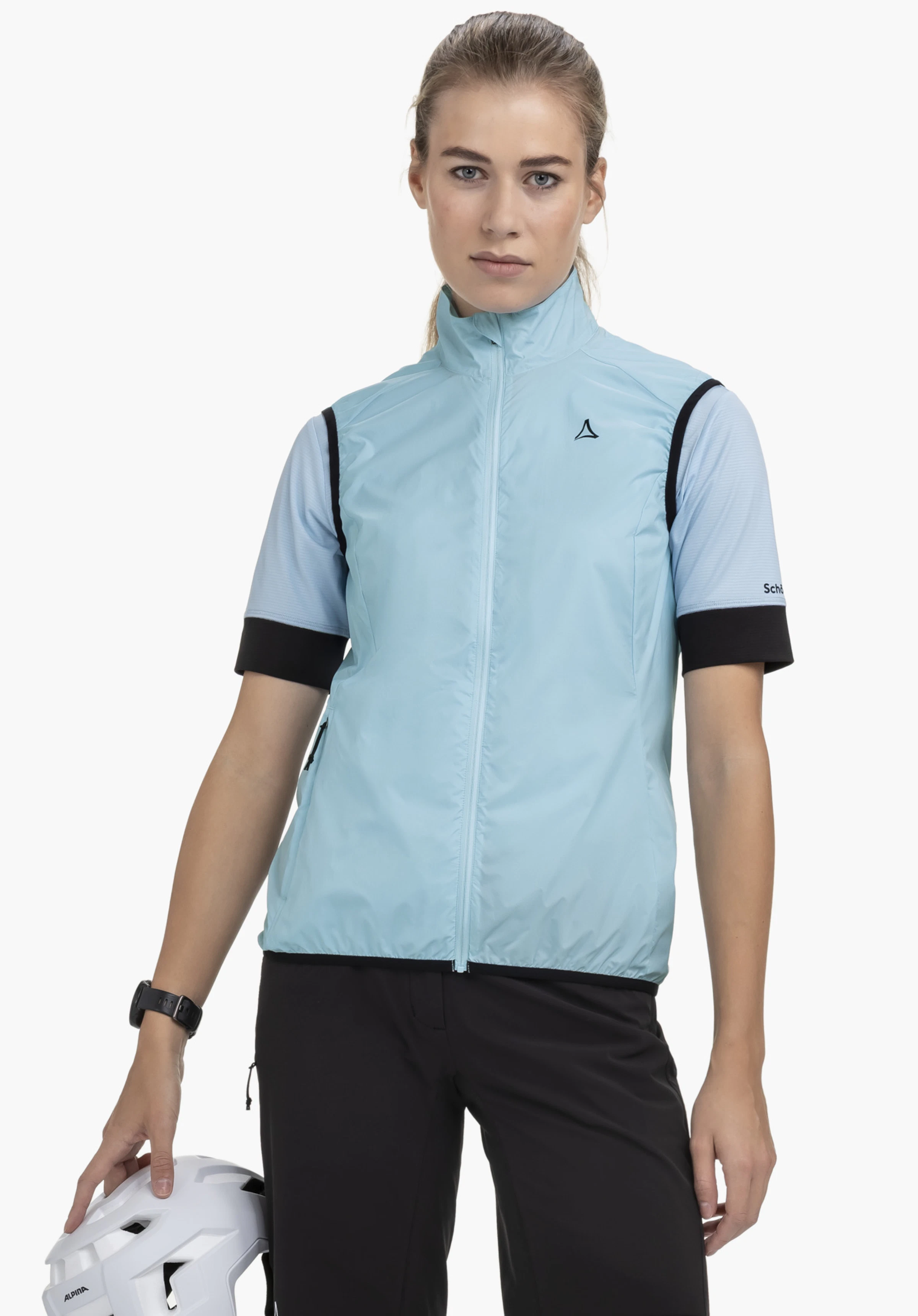 Vest Style Cannobio WMS on a model standing frontally against a neutral background. She is wearing the blue gilet with a visible Schöffel logo on the chest and is holding a bicycle helmet in her hand.