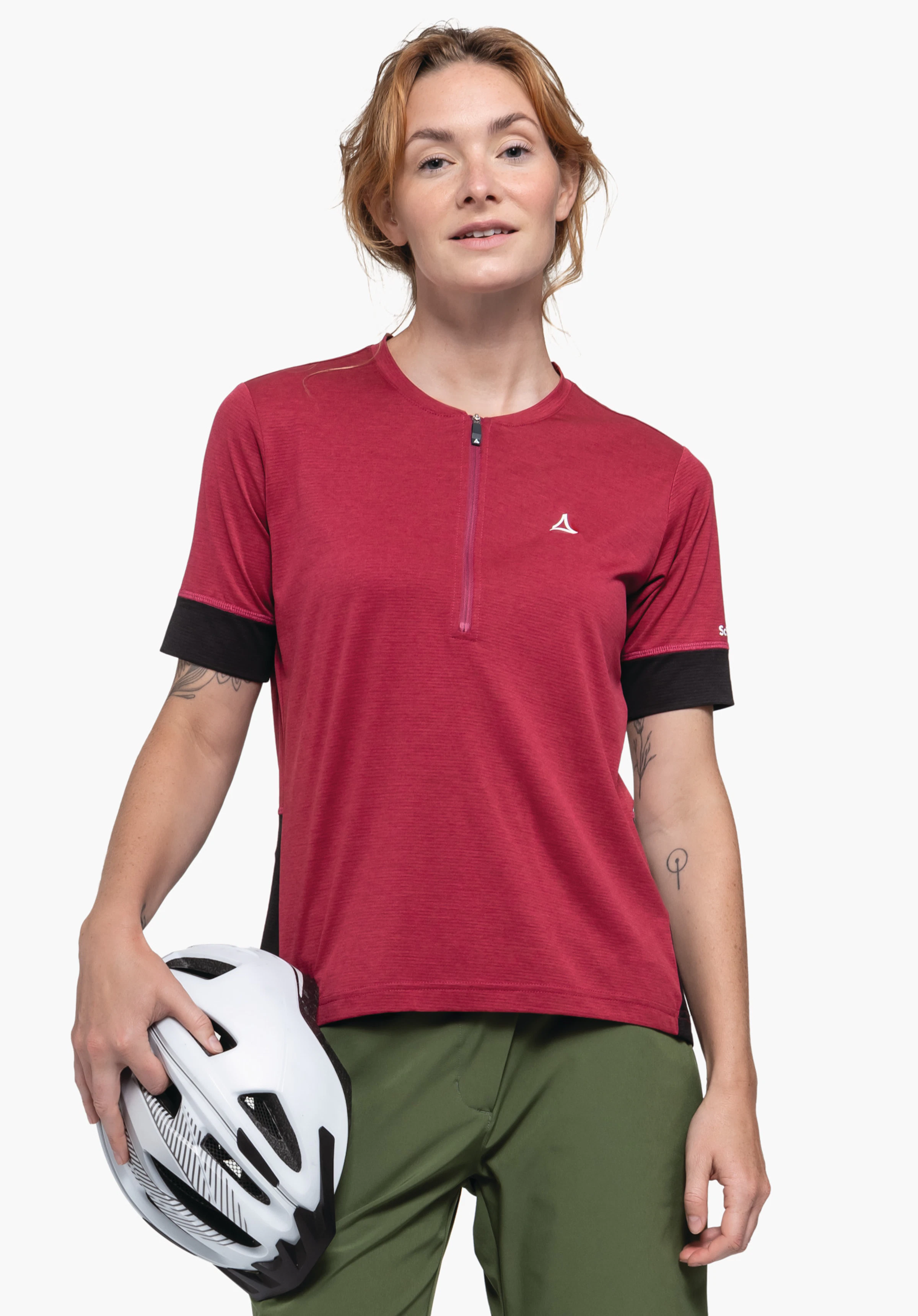 CIRC Shirt Style Pinega WMS femme portant un maillot de cyclisme rouge à manches courtes avec logo Schöffel et un pantalon vert, tenant un casque de vélo blanc; debout devant un fond blanc. Texte sur la manche:Schöffel.