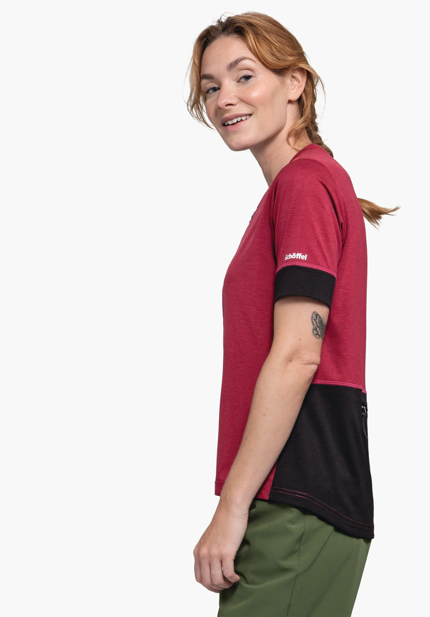 CIRC Shirt Style Pinega WMS femme debout de profil et souriante, porte un t-shirt rouge foncé avec empiècement noir et pantalon vert devant fond clair. Texte sur la manche: "schöffel".