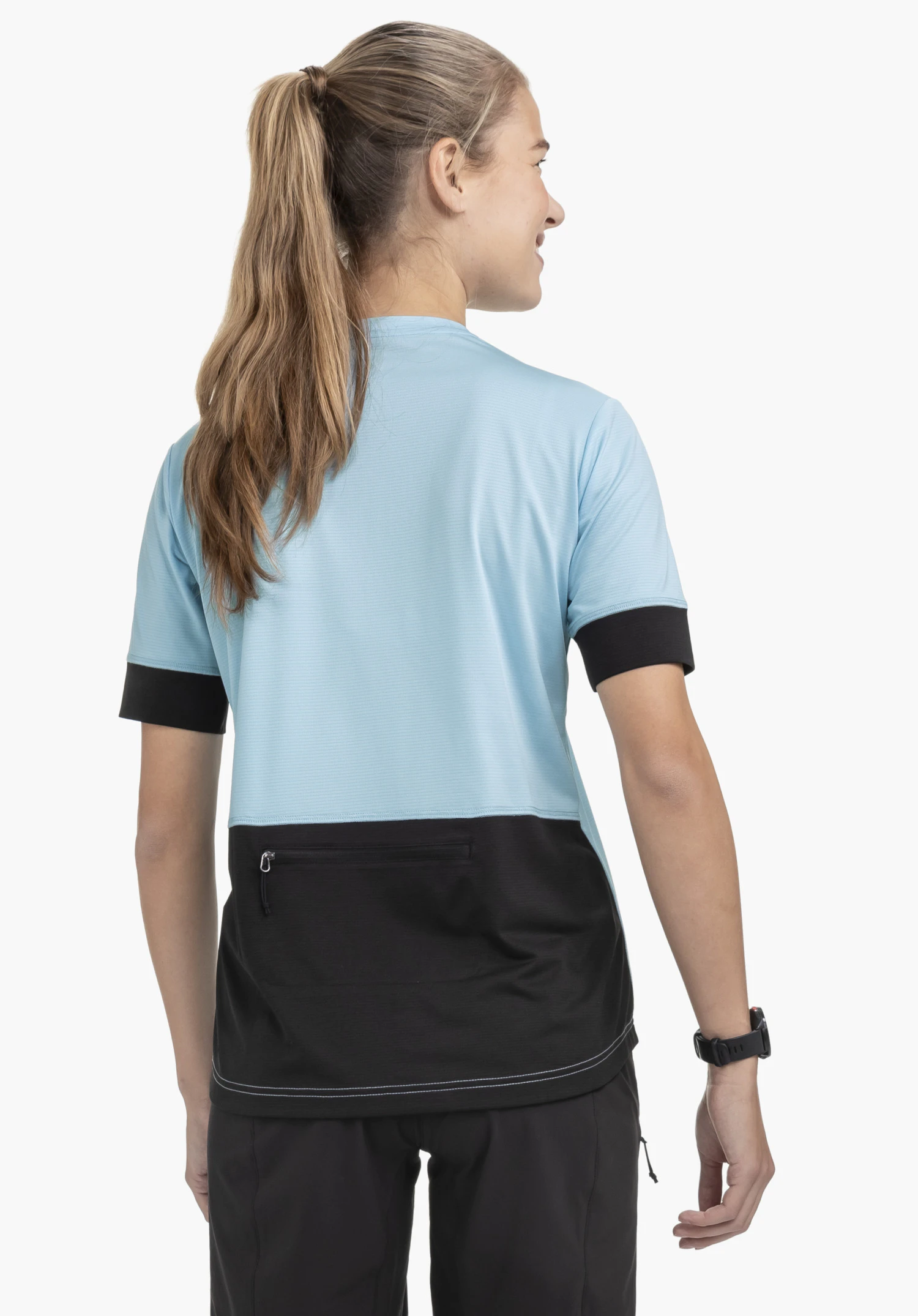 CIRC Shirt Style Pinega WMS en bleu. Une jeune femme est présentée de dos sur un fond neutre, mettant en avant la poche zippée pratique au dos et les bords de manches sombres.