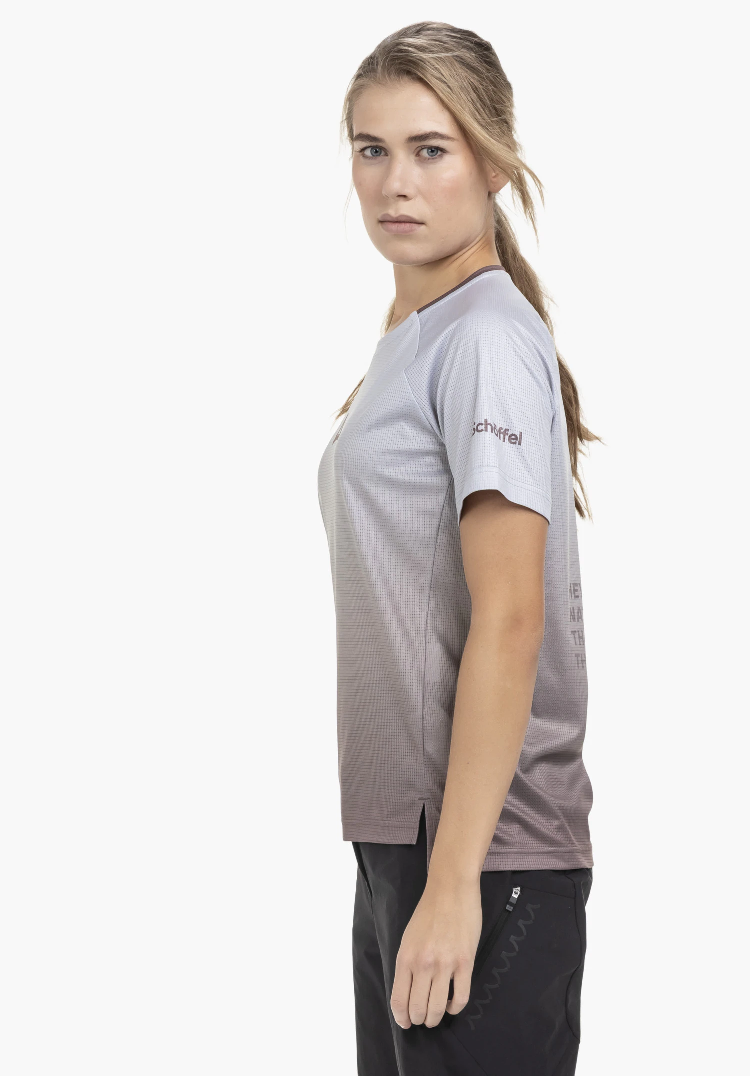 Shirt Style Koitere WMN in braun. Die Sportlerin steht seitlich vor einem neutralen Hintergrund und trägt das Shirt mit dem gut sichtbaren Schöffel-Logo am Ärmel.