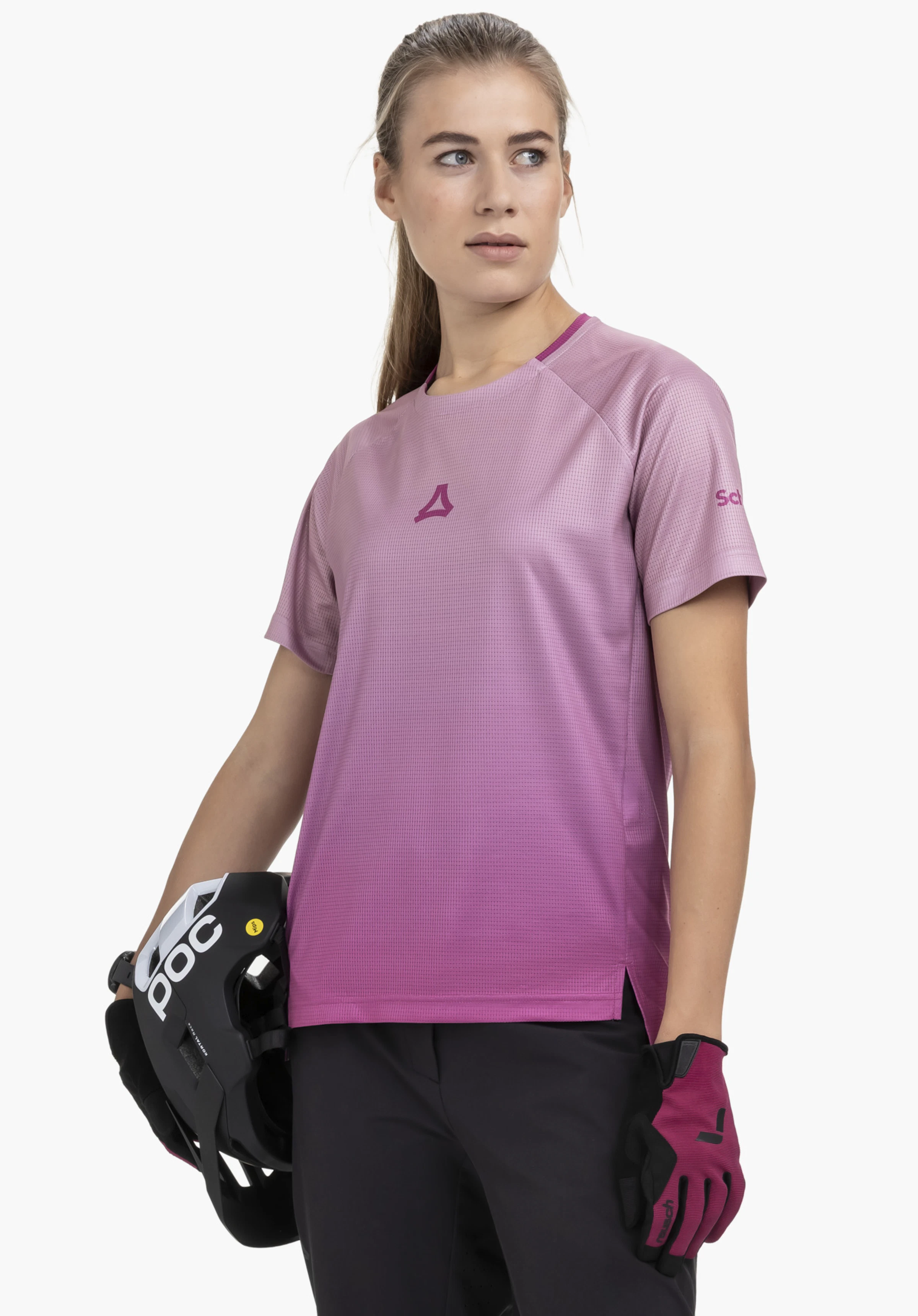 Shirt Style Koitere WMN in pink. The model is standing in a slight half-turn against a neutral background, showcasing the shirt. The subtle Schöffel logo is visible on the chest and the sleeve. The shirt in pink highlights a sporty look while the model holds a helmet in her hand.