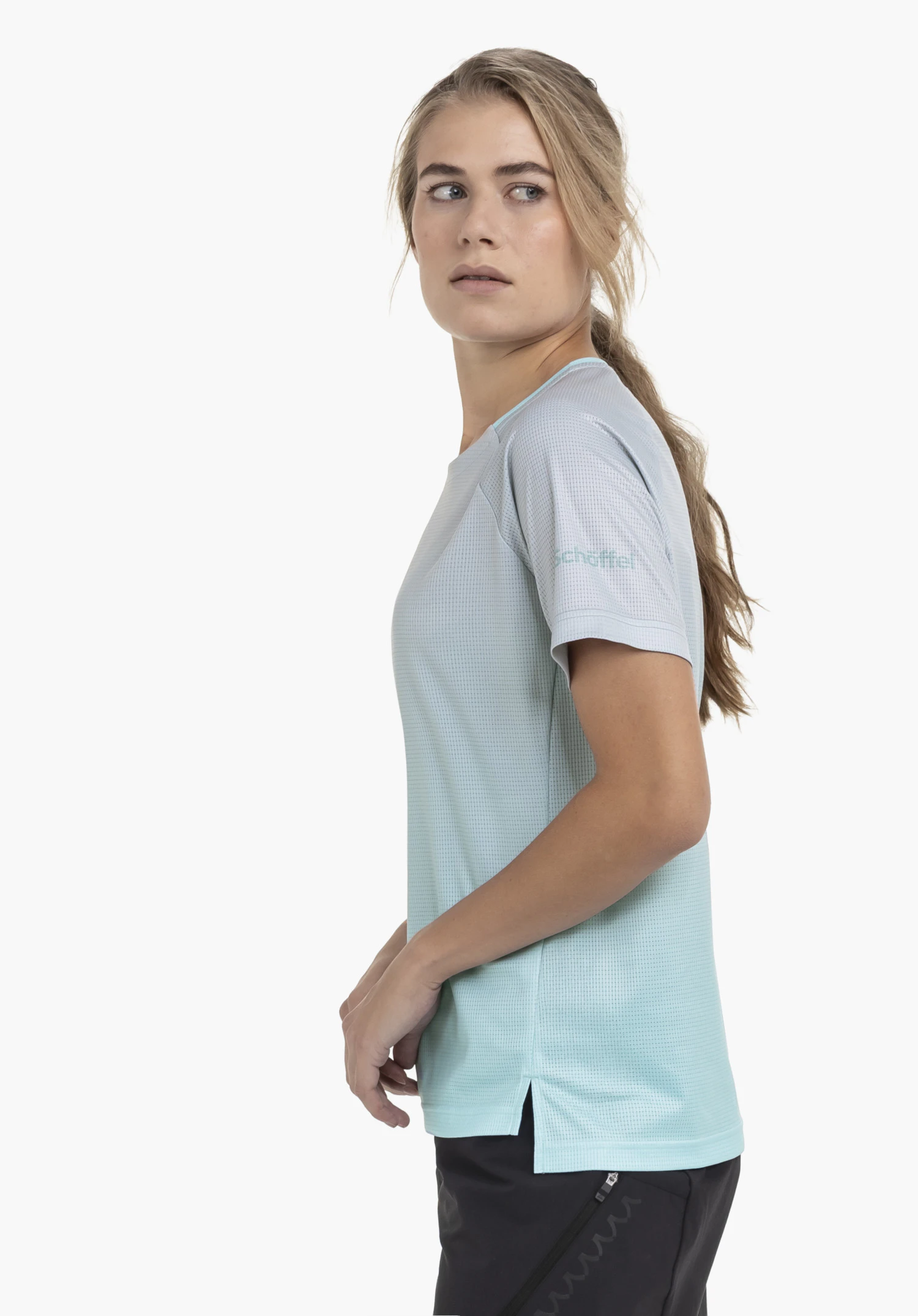 Shirt Style Koitere WMN in Blau: Das Model steht seitlich vor einem neutralen Hintergrund und blickt über die Schulter, wodurch das dezente Schöffel-Logo auf dem Ärmel gut zur Geltung kommt.