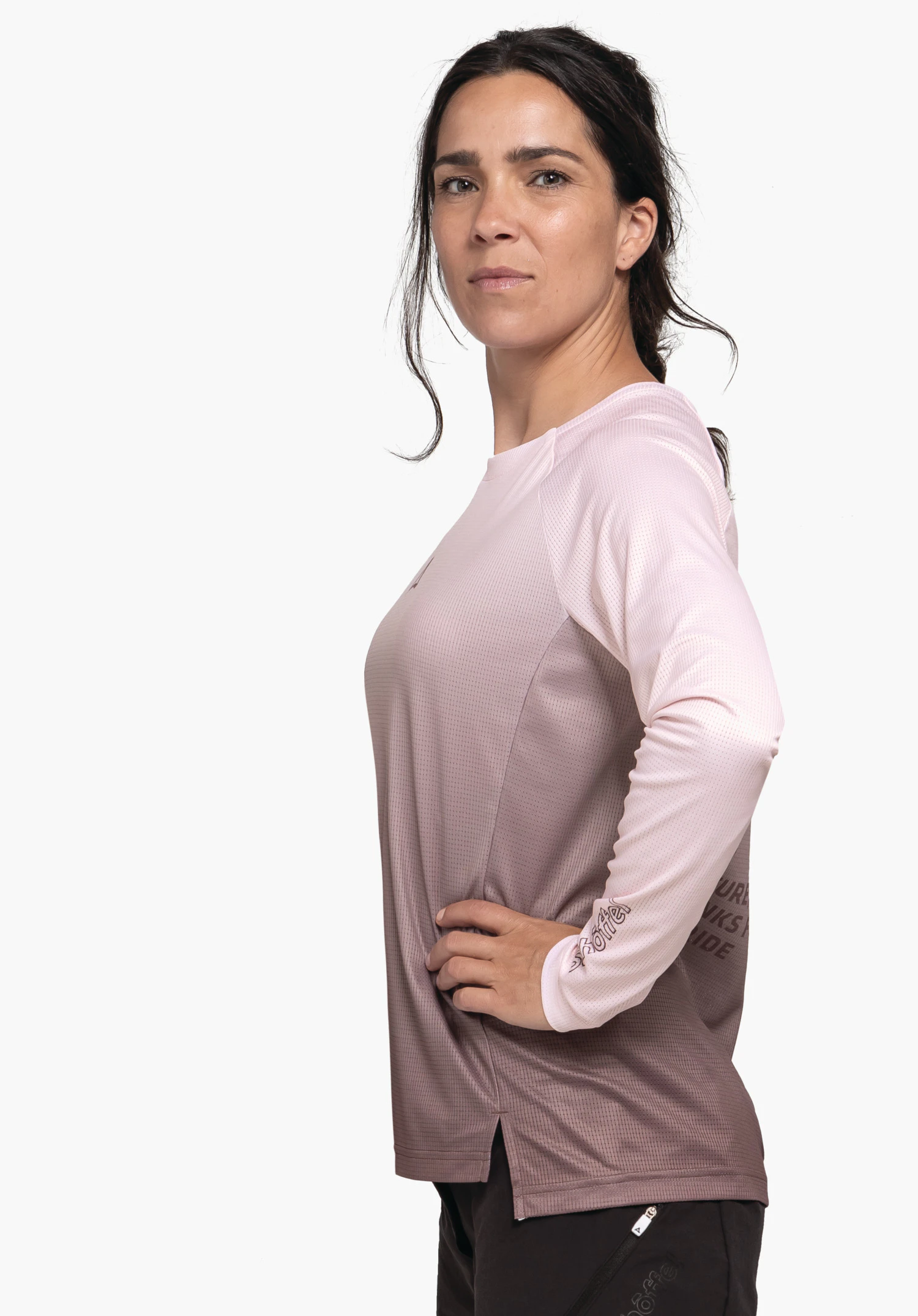Longsleeve Style Koitere WMS personne portant un t-shirt manches longues rose clair, debout de profil, main sur la hanche, pantalon noir; cheveux brun foncé attachés; fond blanc.