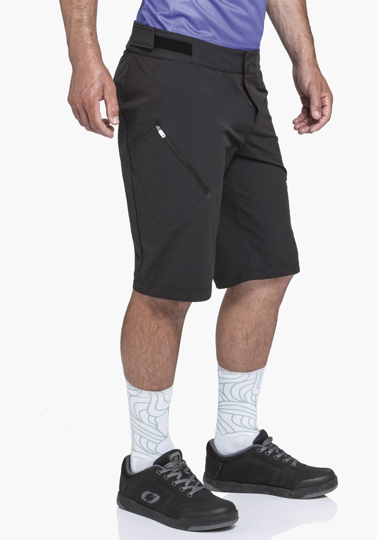 Shorts Style Puula MNS black shorts worn by a man; stands with legs slightly apart; light lilac top, white socks with light green pattern and black shoes; white background.