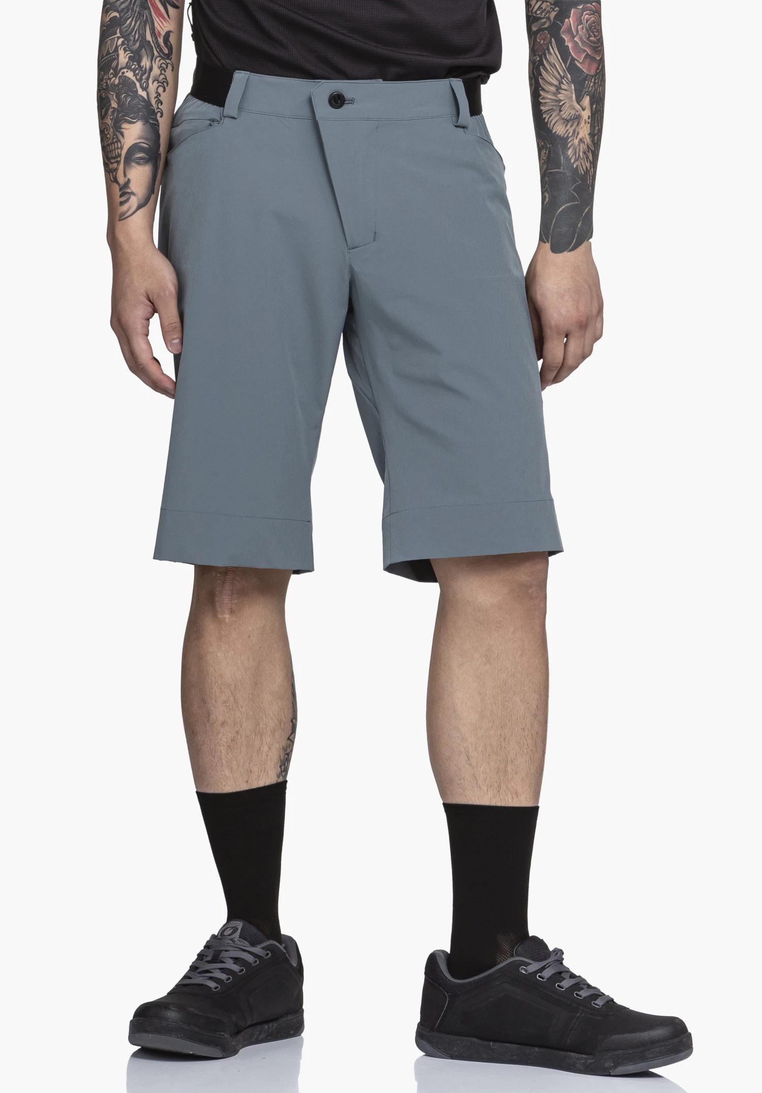 Shorts Style Keitele MNS donkerblauwe shorts, gedragen door een staande persoon voor een witte achtergrond; knielang, zwarte sokken en schoenen, zichtbare tatoeages op de onderarmen, handen ontspannen langs het lichaam.