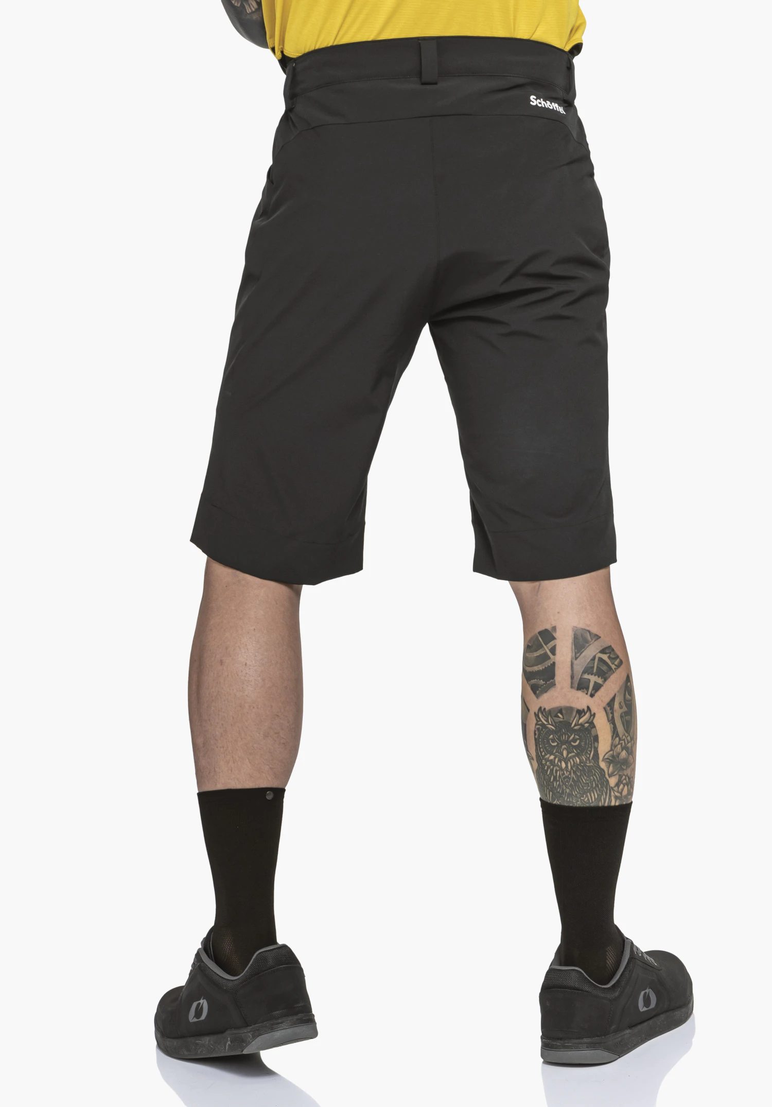 Shorts Style Keitele MNS black shorts worn by a standing person with a light yellow top; legs slightly apart against a white background; “Schöffel” logo visible on the right side; tattoo on the right calf.