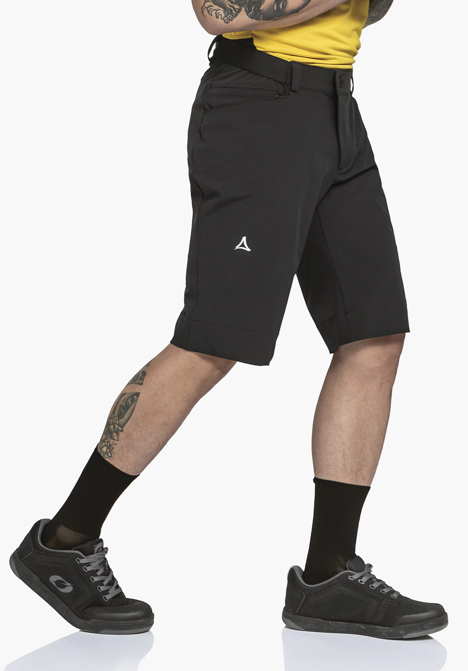 Shorts Style Keitele MNS black shorts with Schöffel logo, worn by a person taking a step; light yellow shirt, tattooed leg, black socks and black shoes in front of a white background.