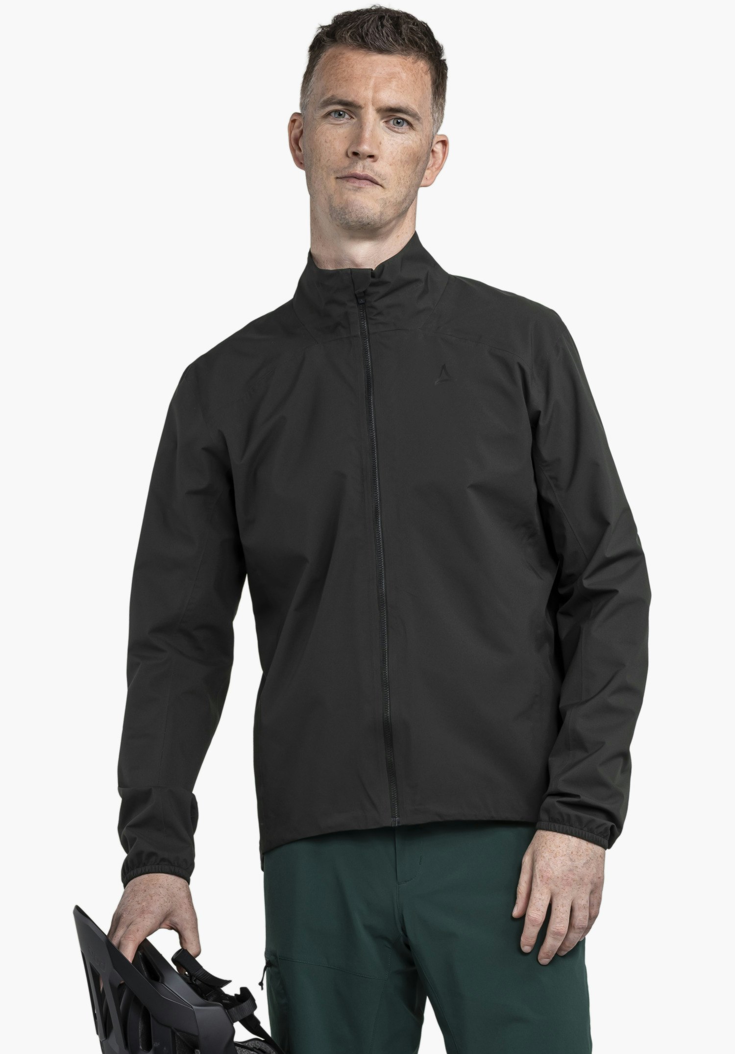 Jacket Style Lurbek MNS worn by a model standing frontally and holding a helmet in his hand. The black jacket is fully zipped and features a subtle Schöffel logo on the left chest, presented against a neutral background.
