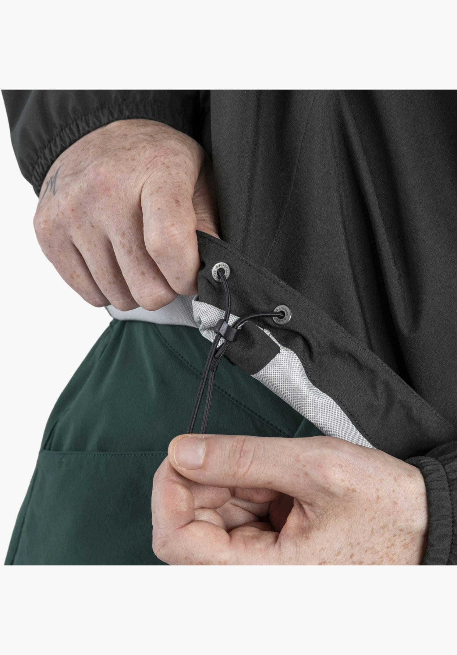 Jacket Style Lurbek MNS shows the adjustable hem in black in detail. Hands are adjusting the elasticated drawcord with a toggle, revealing the light inner lining and metal eyelets featuring the Schöffel-Logo, close-up in front of a white/neutral background.