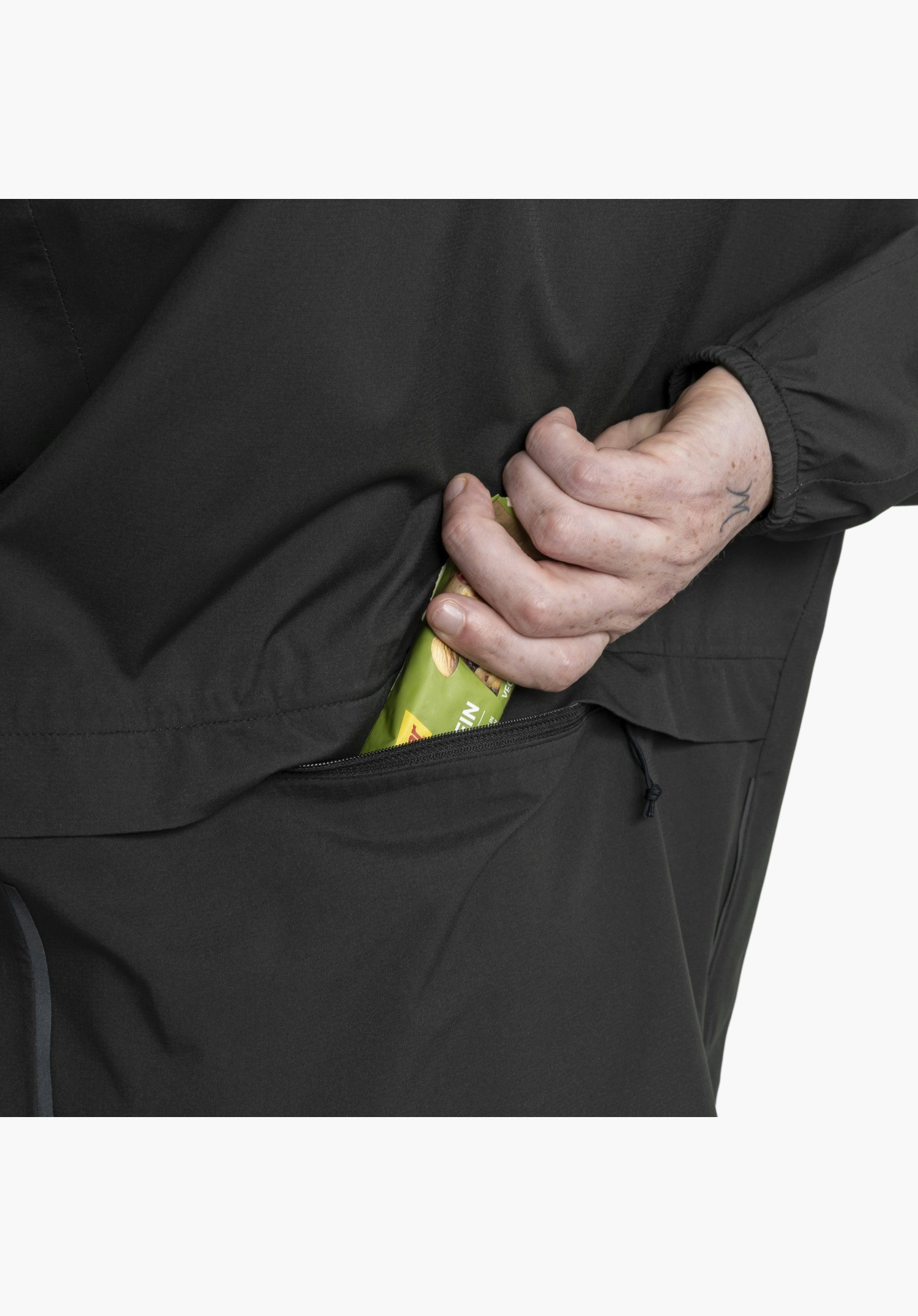 Jacket Style Lurbek MNS shows the practical, concealed zip pocket in the black fabric, from which a hand is pulling out a snack bar. Close-up against a white background.