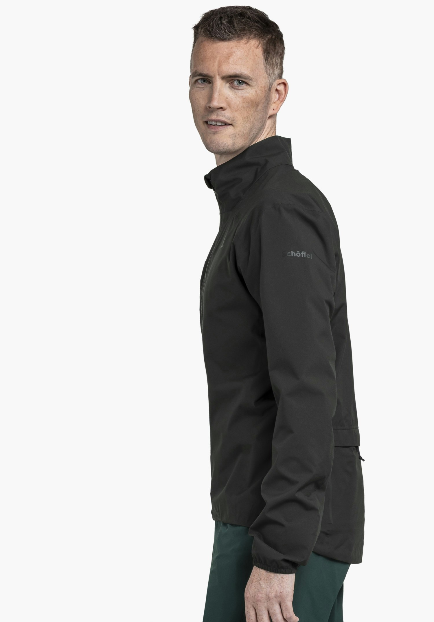 Jacket Style Lurbek MNS in black, worn by a model standing in a side profile pose against a neutral background. The subtle Schöffel logo on the upper arm perfectly complements the clean aesthetic of this functional jacket.