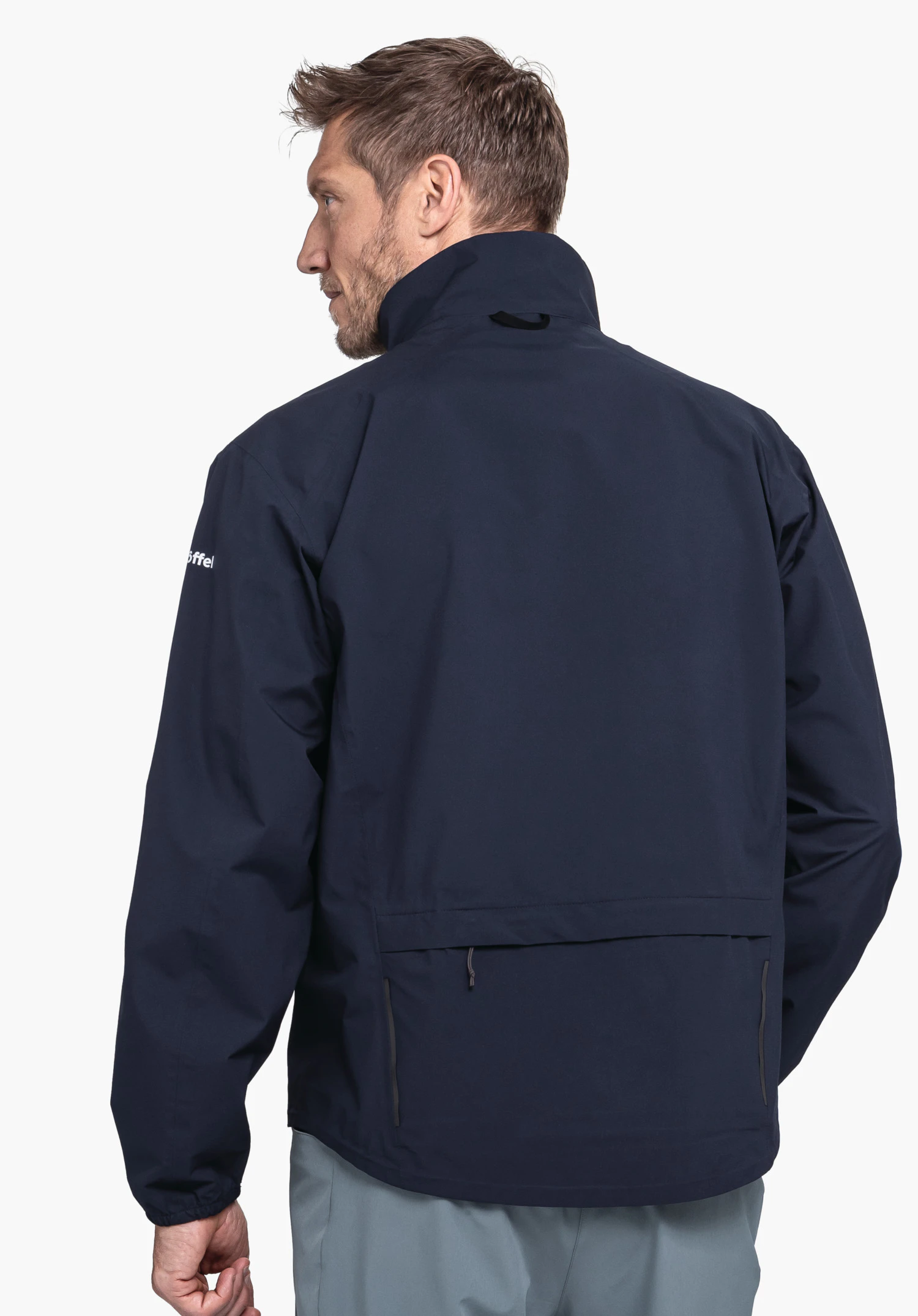 Jacket Style Lurbek MNS dunkelblaue Jacke, getragen von einem Mann, der den Rücken dem Betrachter zukehrt; steht vor weißem Hintergrund; Ärmel mit weißer Aufschrift "Schöffel"; Hose grau hell.