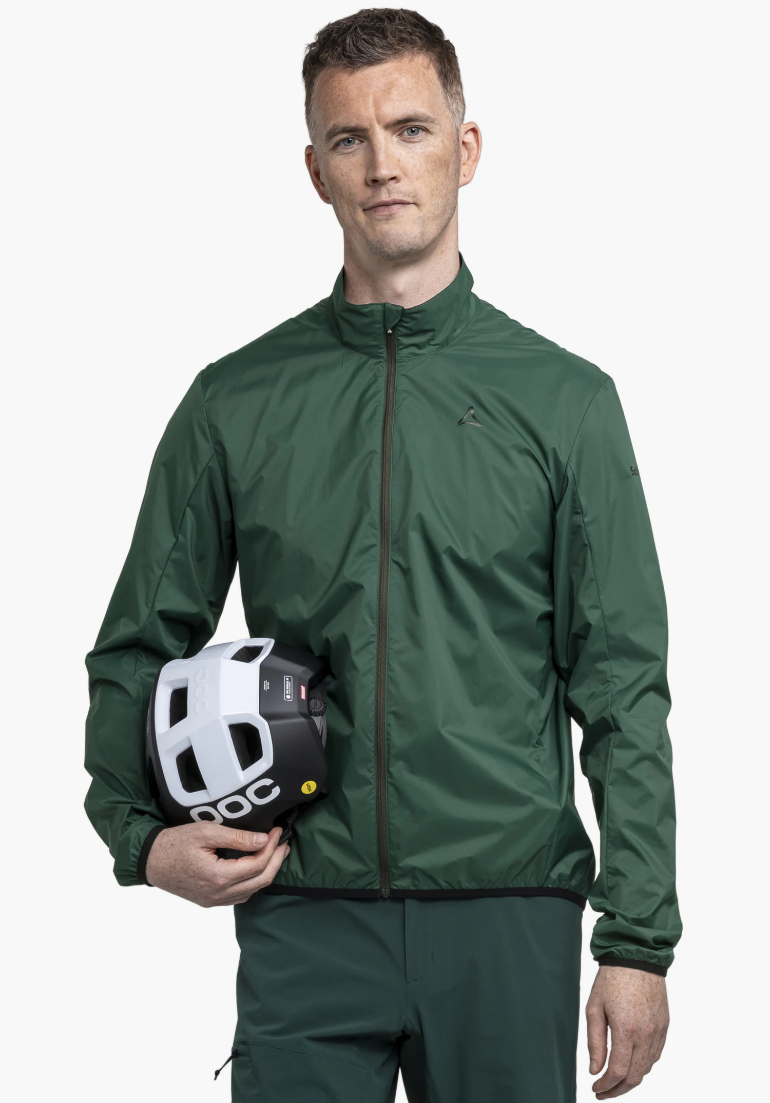 Jacket Style Cannobio MNS. A model stands facing forward in front of a neutral background, presenting the jacket in green. The Schöffel logo is visible on the left chest, while the man holds a bicycle helmet under his arm, emphasising its sporty intended use.