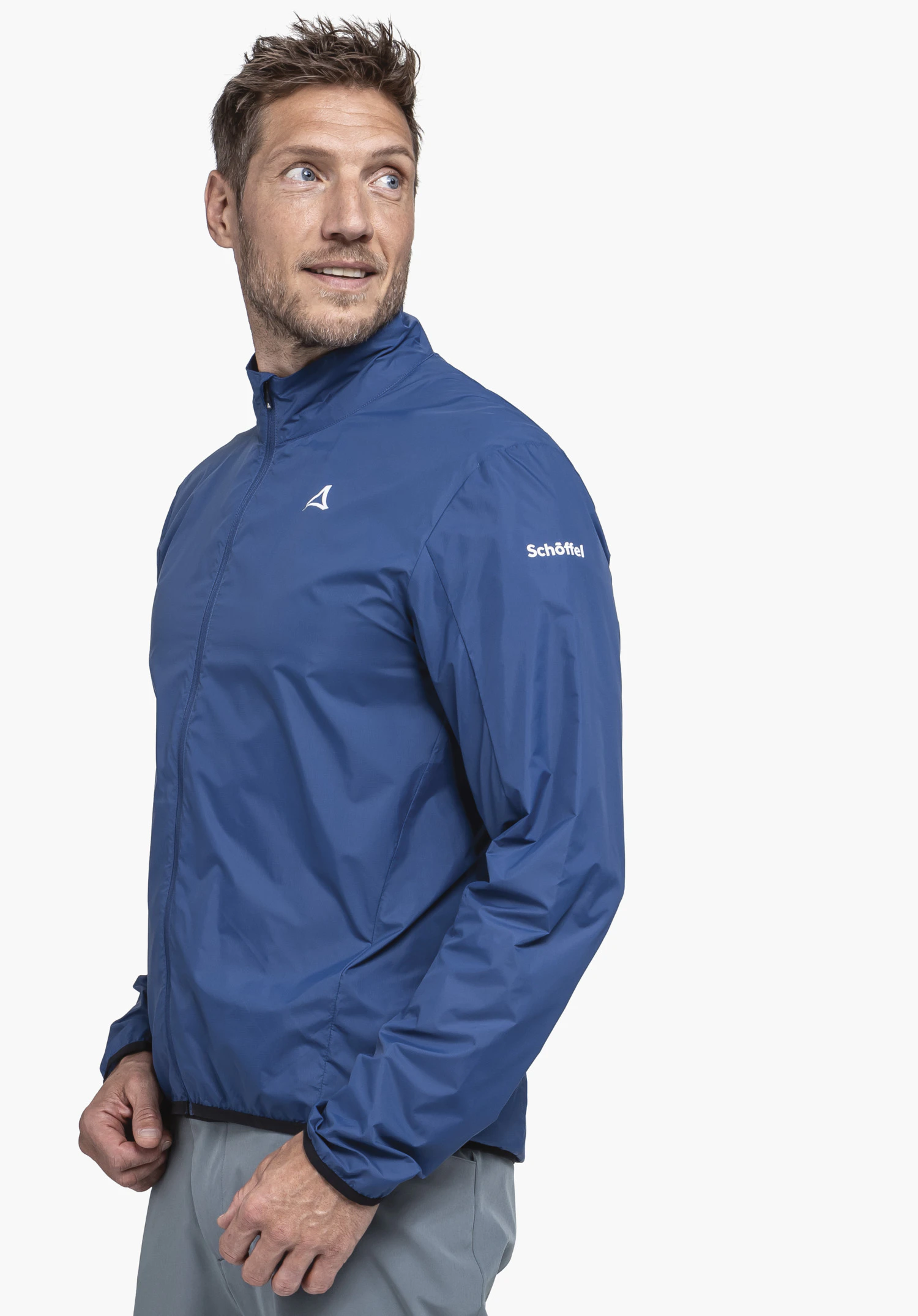 Jacket Style Cannobio MNS homme portant une veste coupe-vent bleu foncé avec logo Schöffel sur la poitrine, regarde de côté; fond studio blanc. Texte sur la manche: "Schöffel".