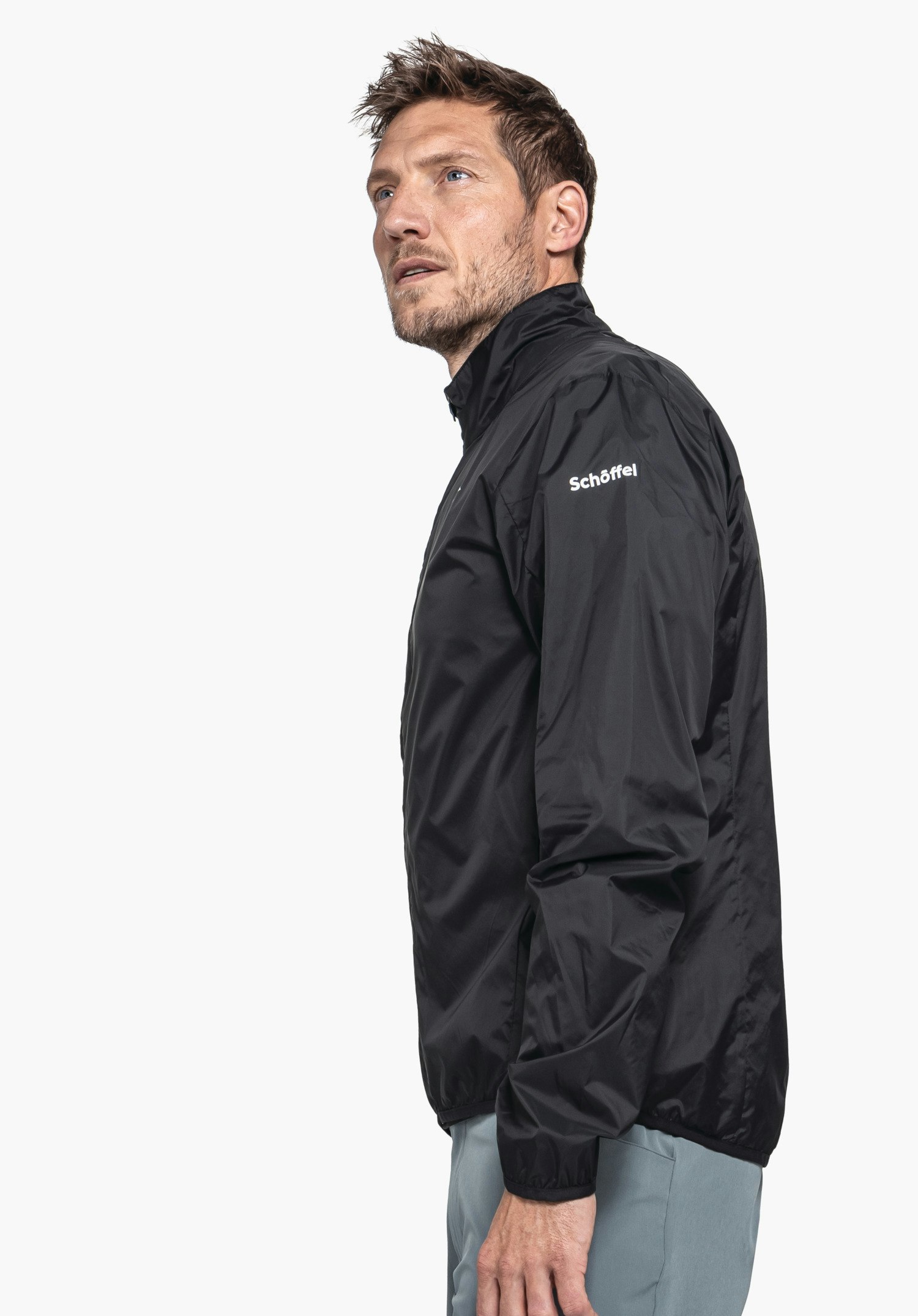 Jacket Style Cannobio MNS Schwarze Jacke, von einem stehenden Mann seitlich getragen, der nach oben blickt; neutraler weißer Hintergrund. Text auf dem Ärmel: "Schöffel".
