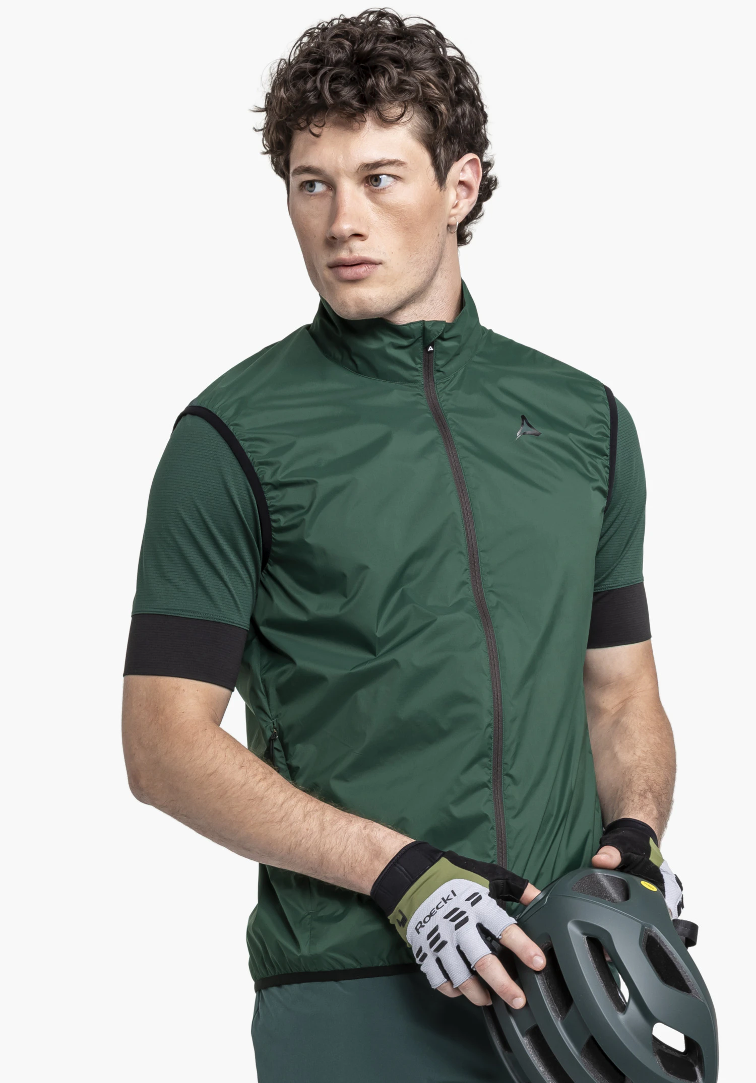 Vest Style Cannobio MNS on a model standing slightly turned to the side, holding a cycle helmet. The green vest features the Schöffel logo on the left chest, photographed against a neutral background.