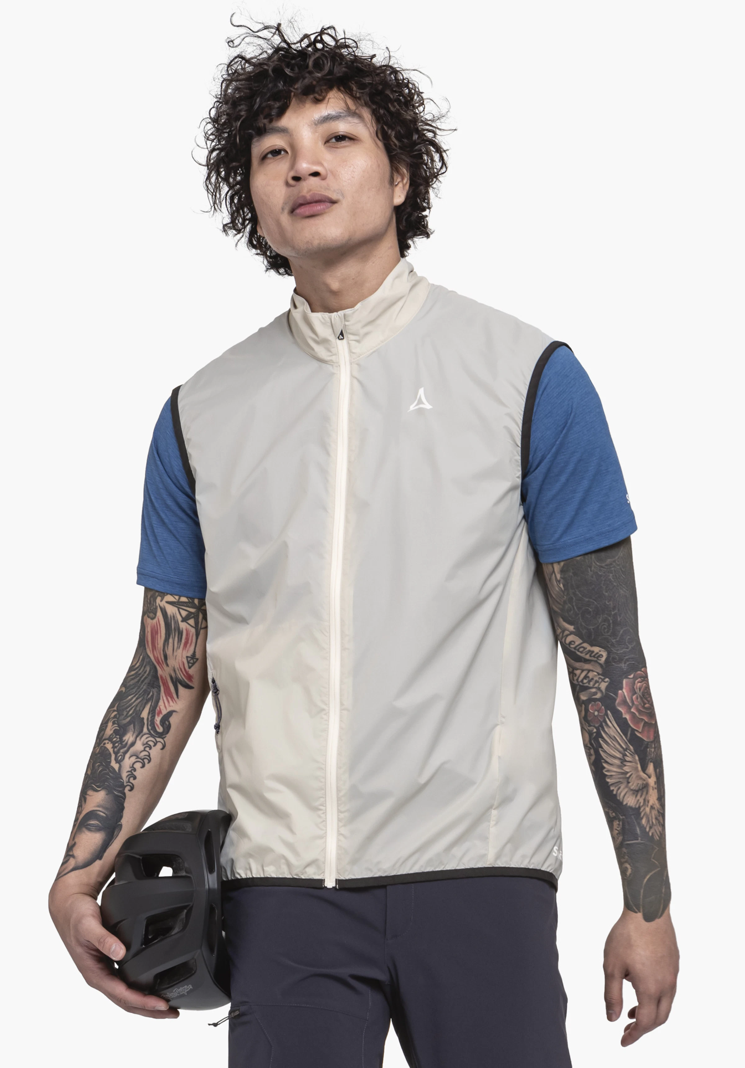 Vest Style Cannobio MNS an einem Model, das frontal steht und einen Fahrradhelm in der rechten Hand hält. Die Weste in der Farbe Natur ist geschlossen und zeigt das weiße Schöffel-Logo auf der linken Brust. Das Bild wurde vor einem neutralen Hintergrund aufgenommen.